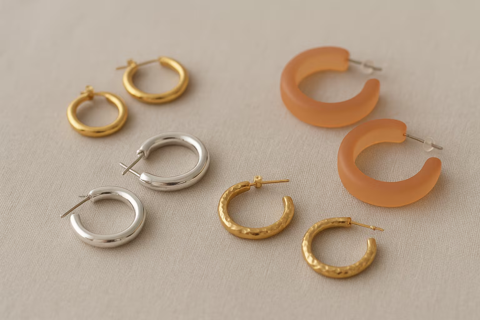 Différentes créoles bijoux posées sur tissu neutre: petites créoles plaquées or, créoles argent, créoles acryliques légères, gros plans sur textures