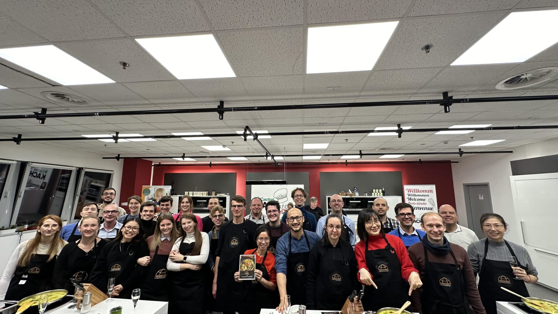 Large corporate cooking class group photo with participants wearing matching aprons in professional kitchen