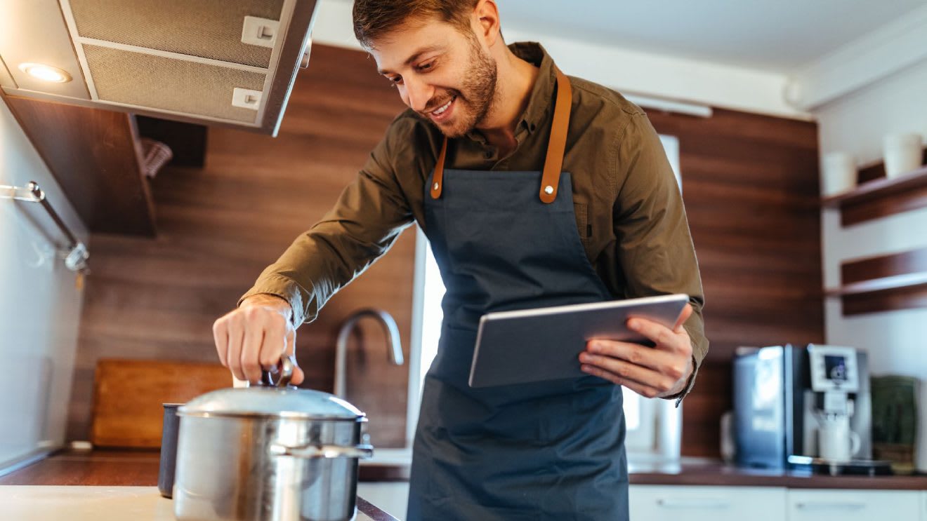 Man in apron cooking while reading tablet recipe in modern kitchen
