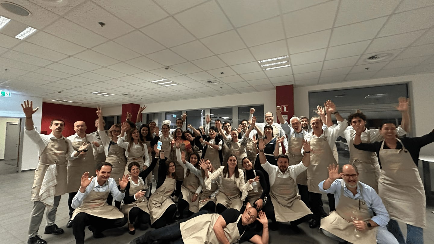 Large culinary team celebrating together in beige aprons waving at camera