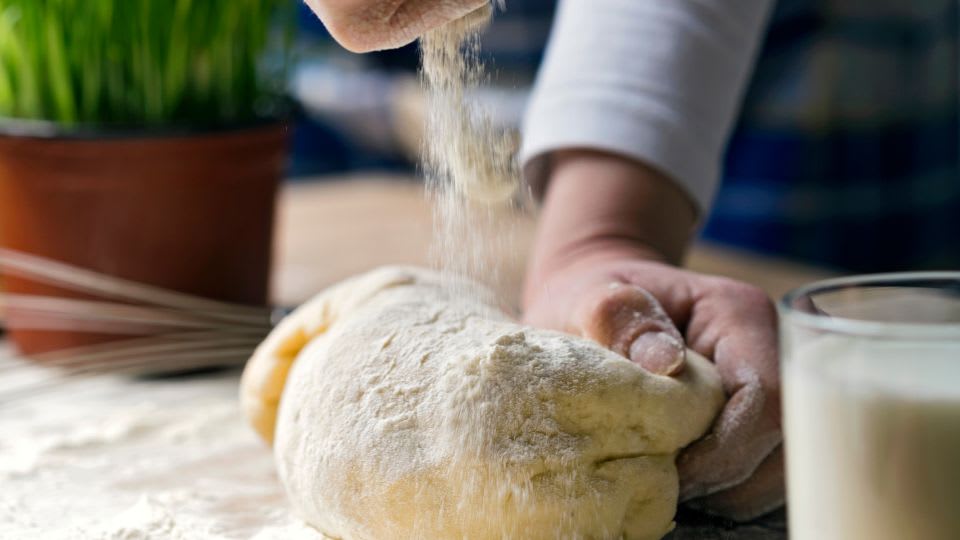 Hands kneading dough with flour falling from above on wooden surface