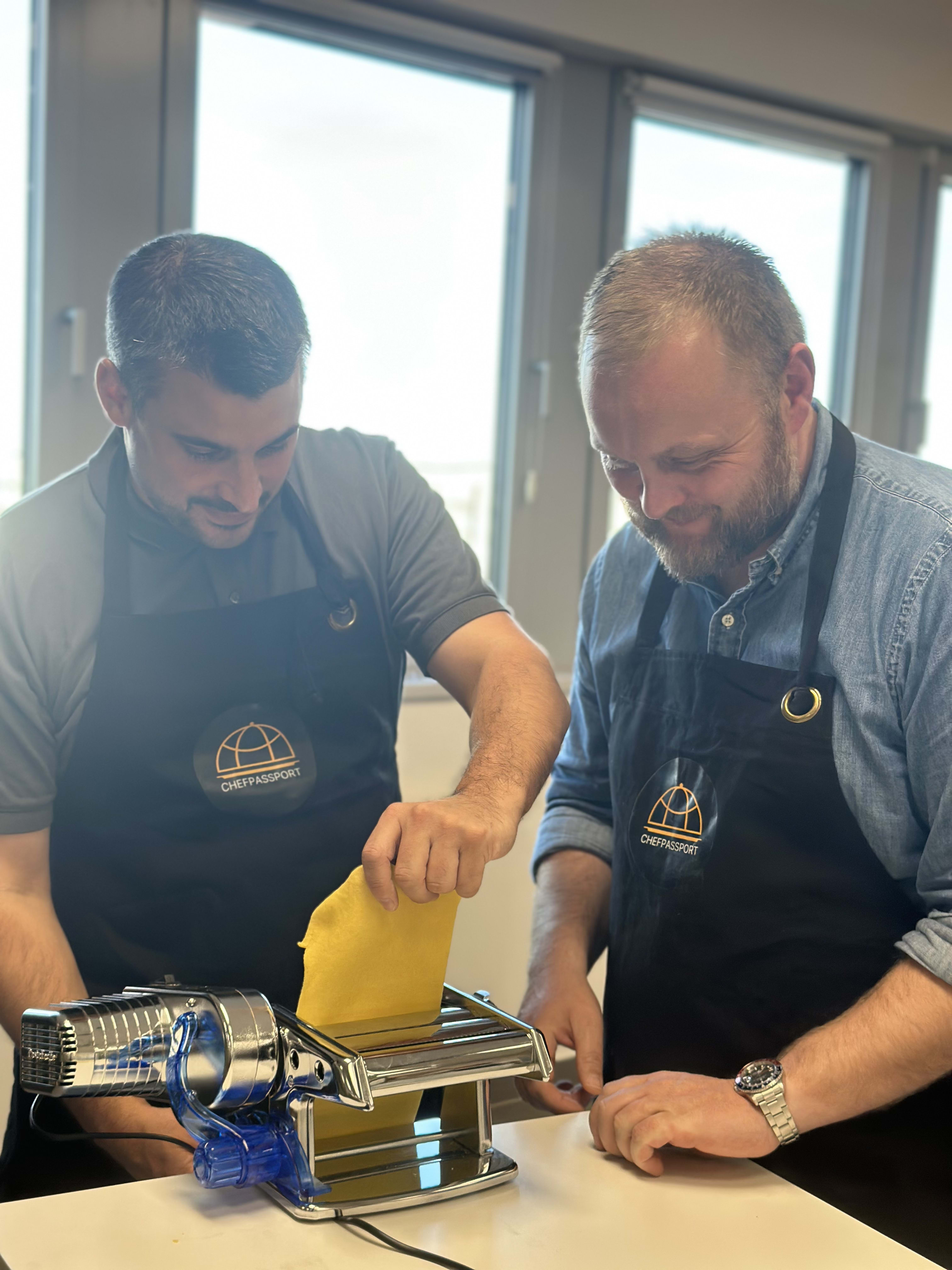 Two chefs in CHEFPASSPORT aprons making fresh pasta with an electric pasta machine