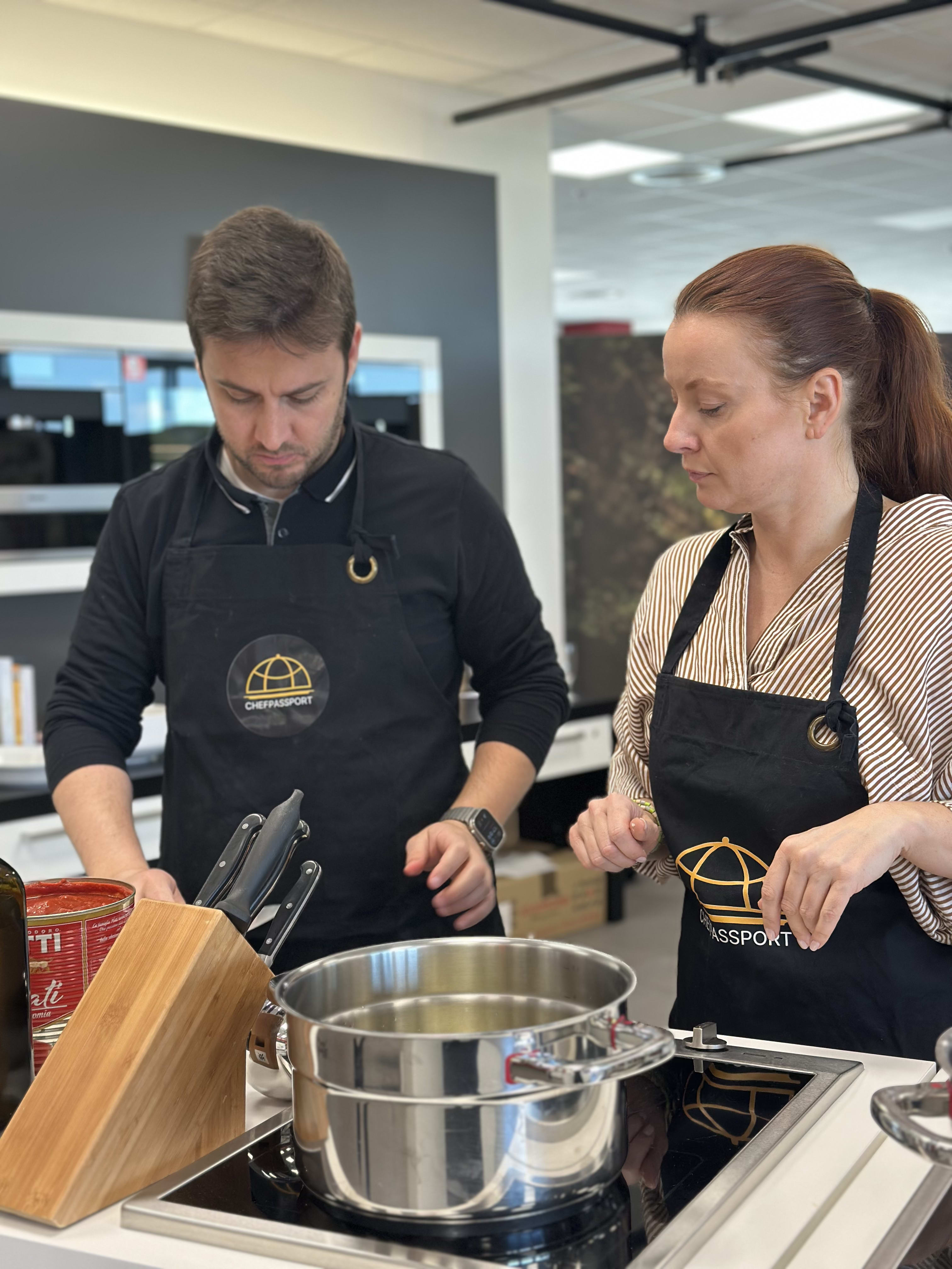 Two people wearing Chefpassport aprons cooking together in a modern kitchen with stainless steel pot