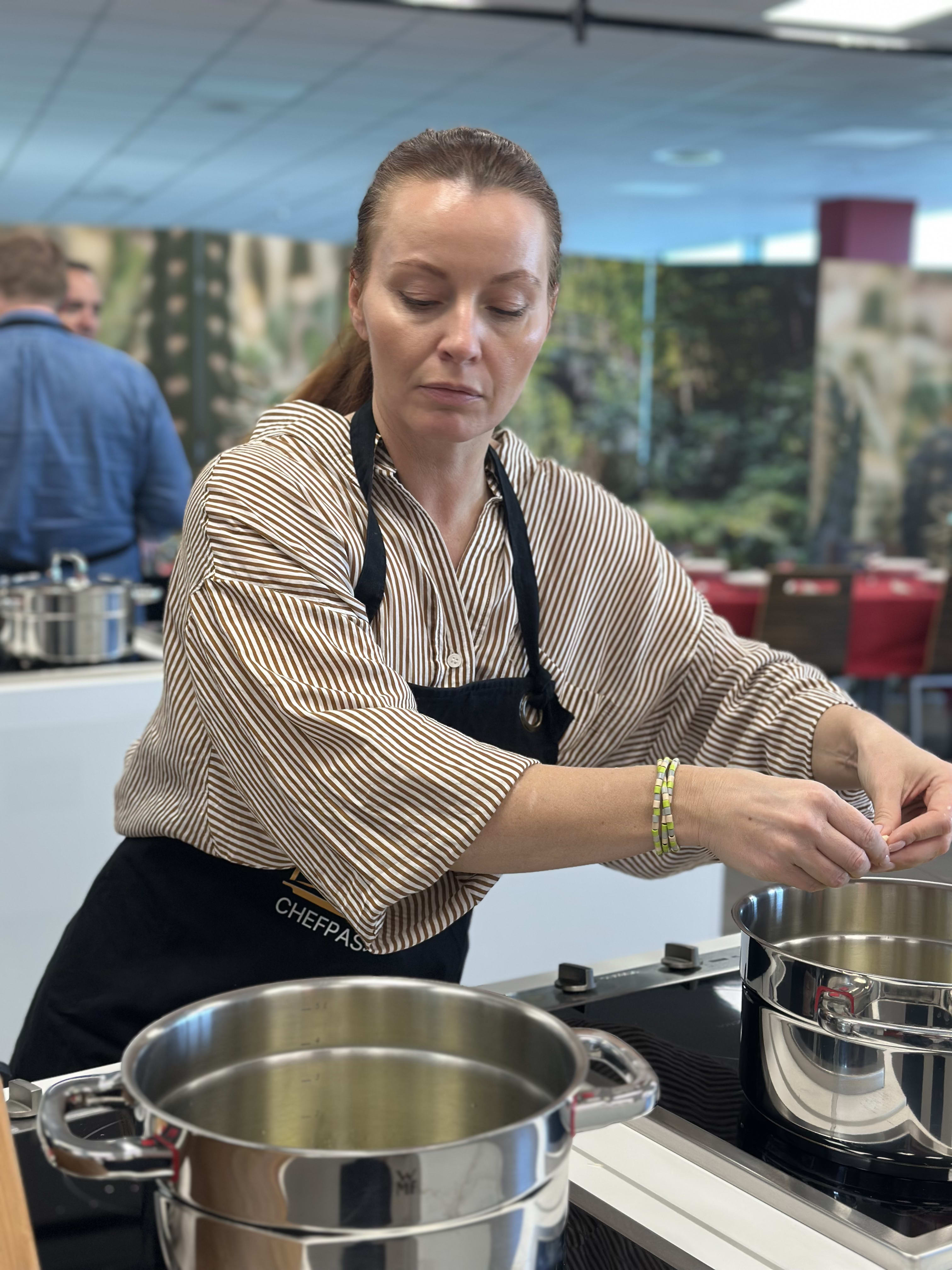 Woman in striped shirt and black apron cooking with stainless steel pots in professional kitchen