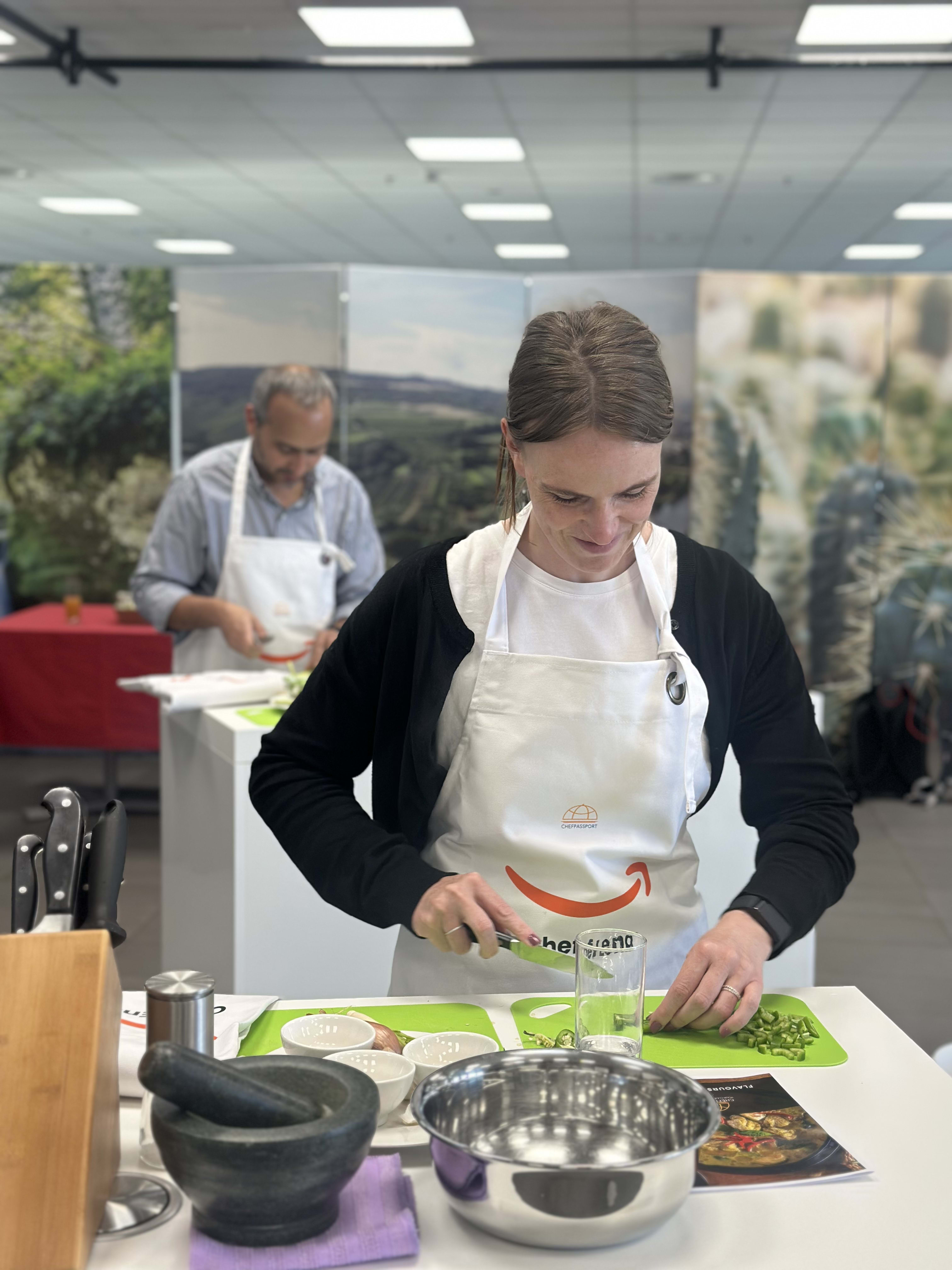 Woman in white apron chopping green vegetables at cooking class with instructor in background