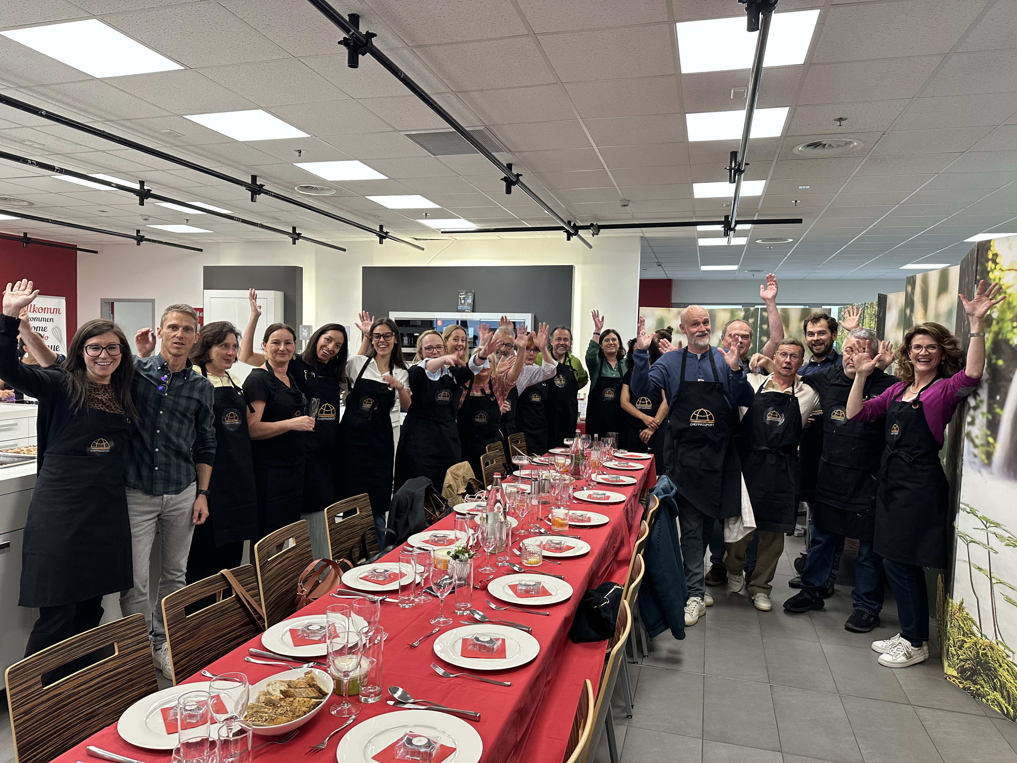 Group of people in black aprons waving hands around long red dining table