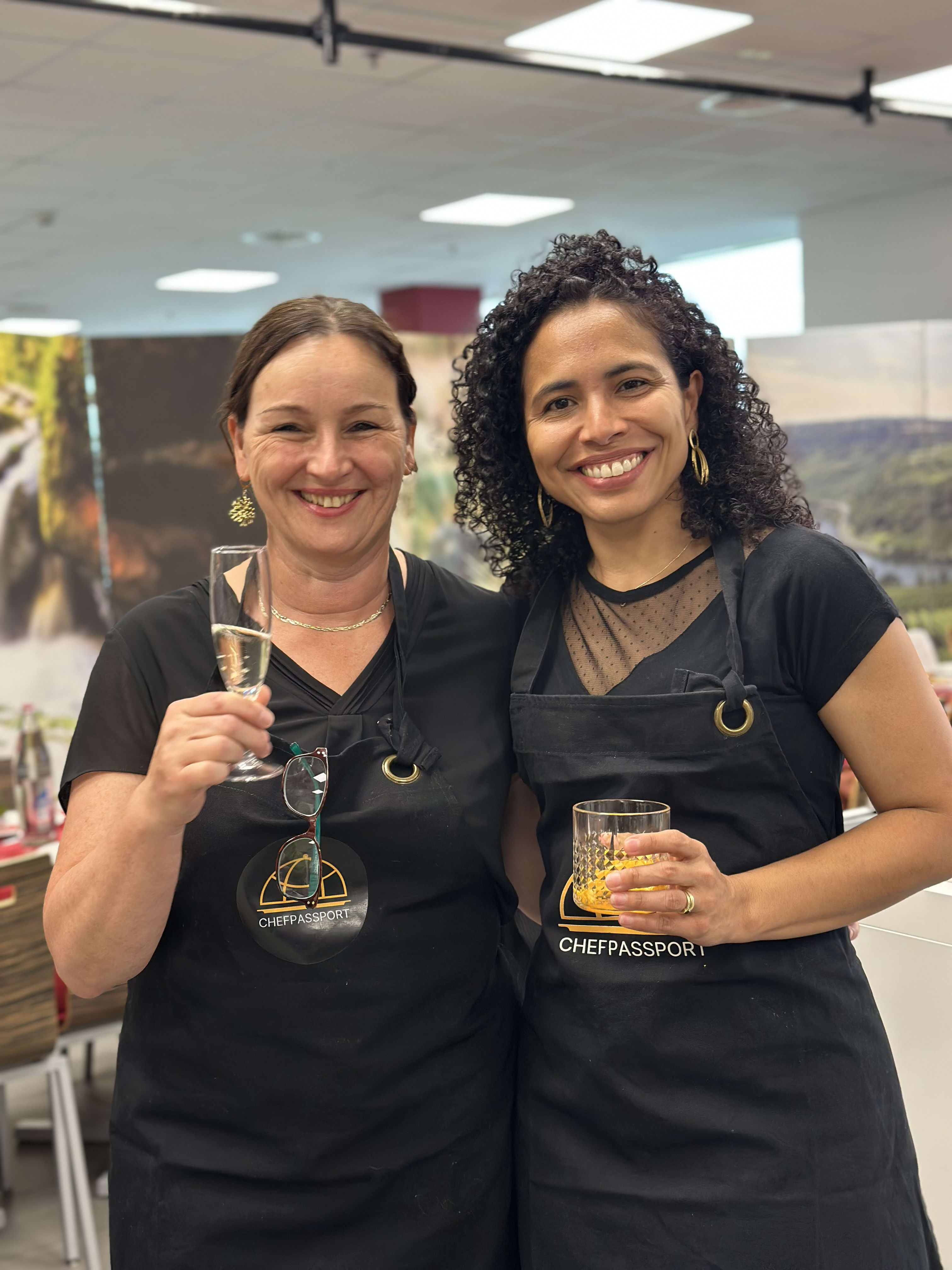 Two women in ChefPassport aprons smiling and holding drinks at a culinary event