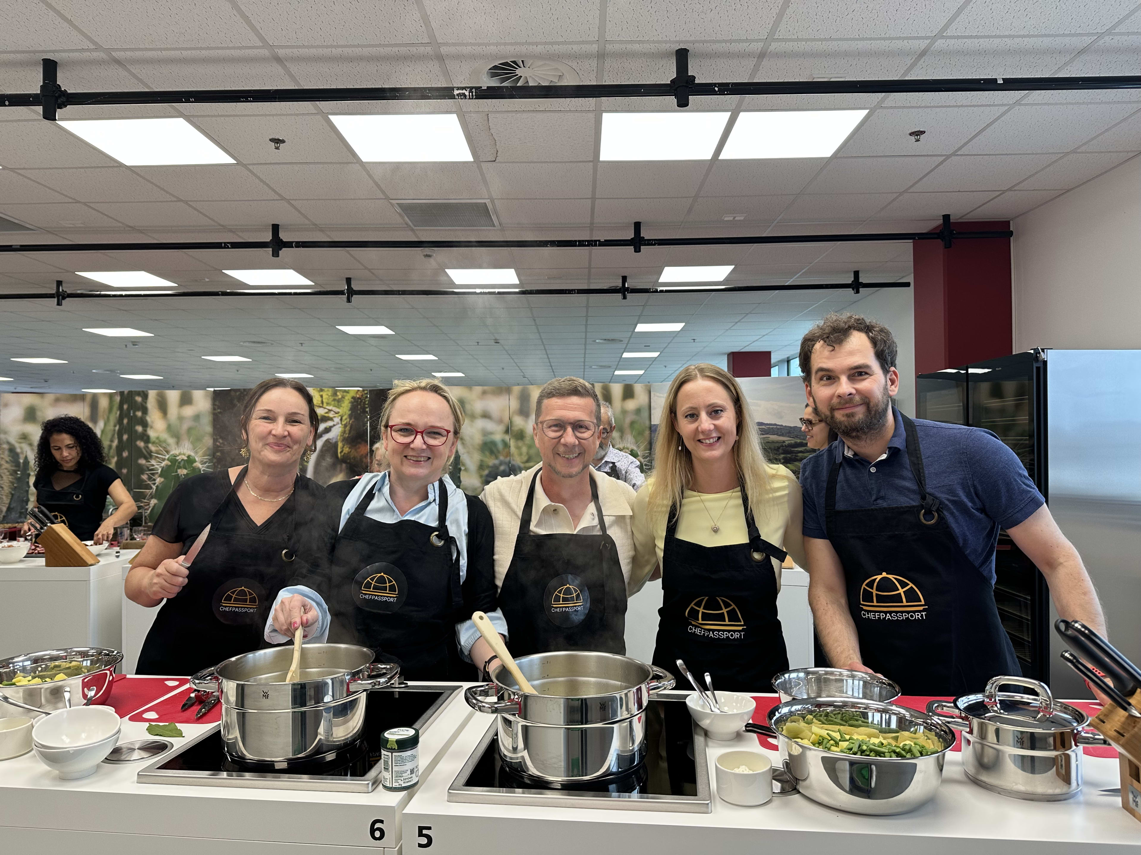 Five people wearing Chef Passport aprons cooking together in a commercial kitchen workspace