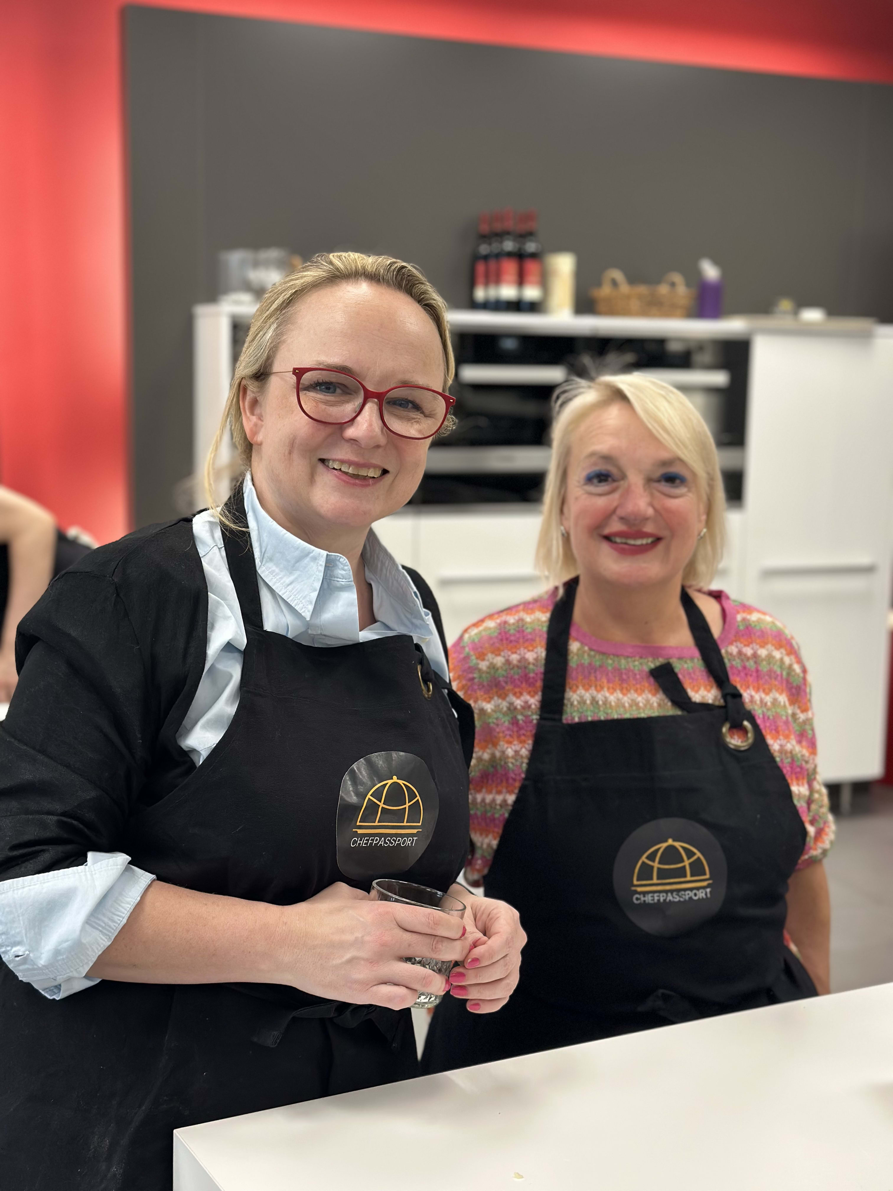 Two women wearing Chef Passport aprons smiling together in a modern kitchen with red walls