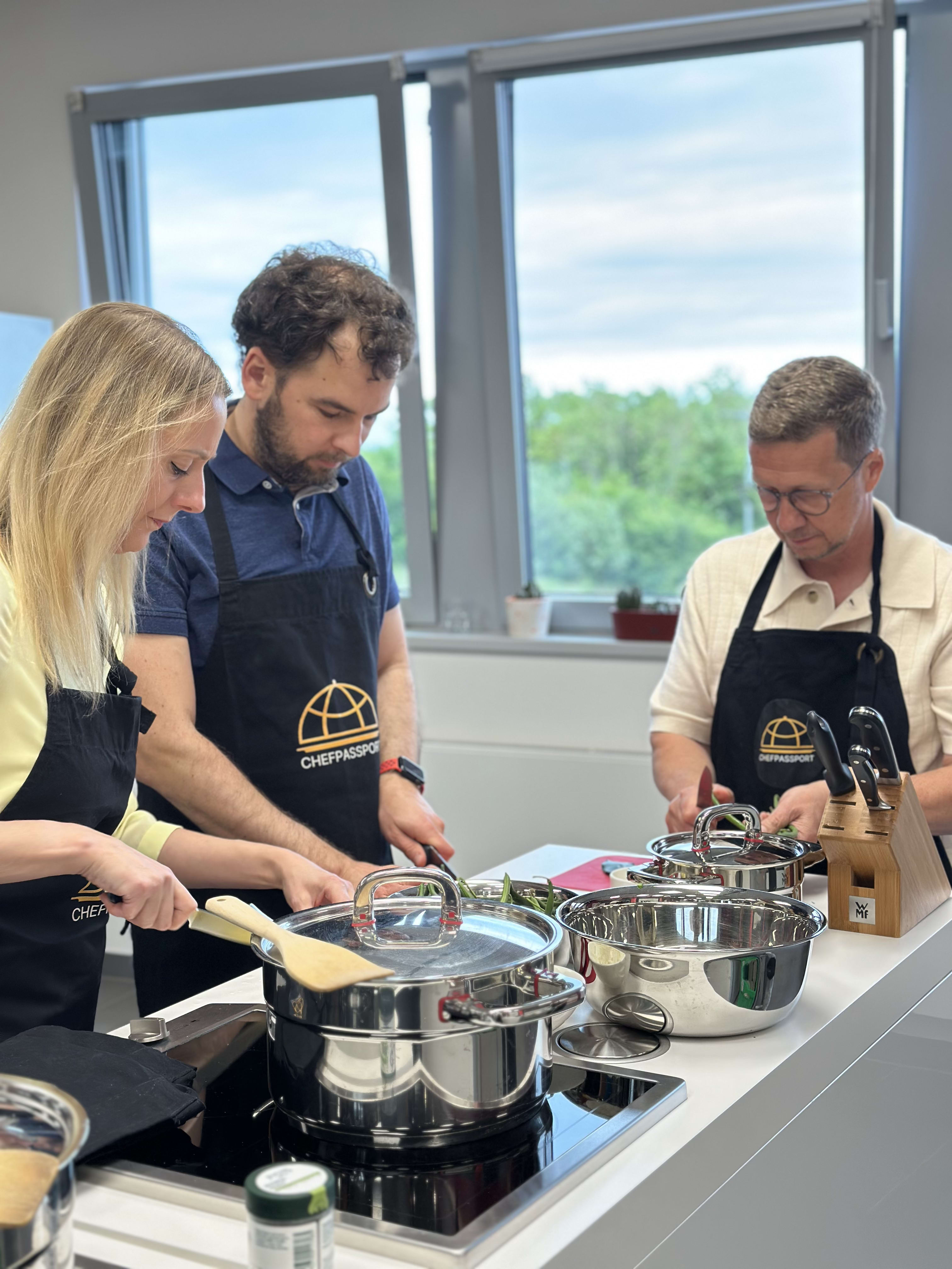 Three people in Chef's Passport aprons cooking together with stainless steel pots in modern kitchen