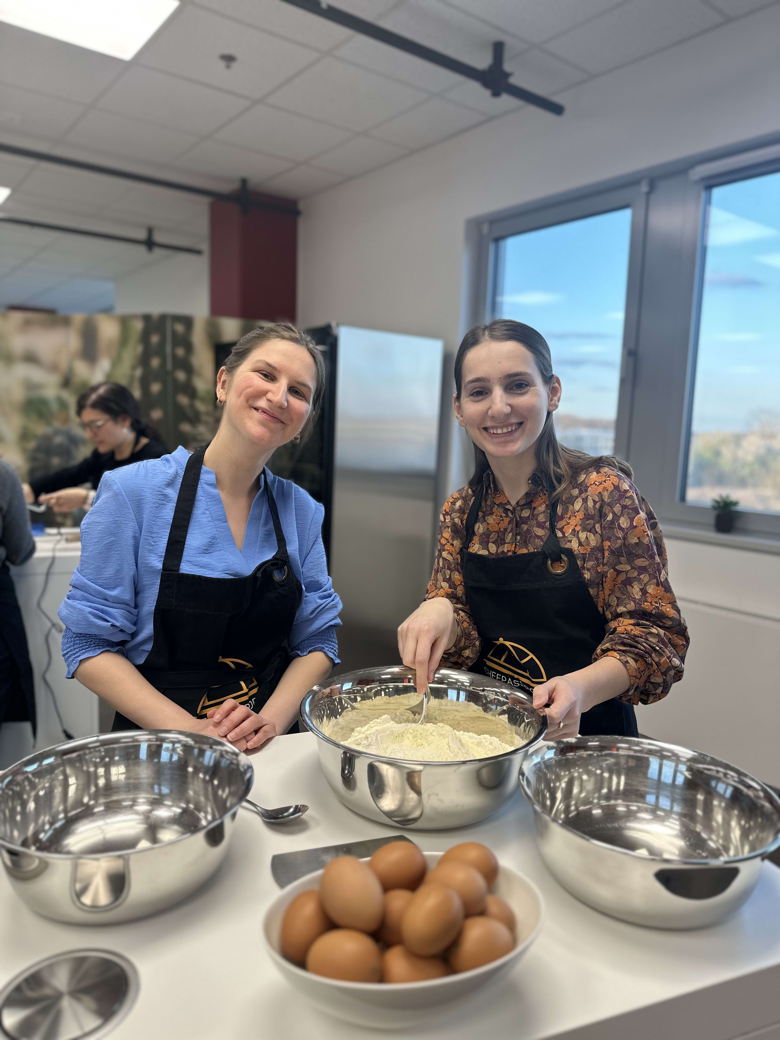 Two women in black aprons preparing food with eggs and flour in stainless steel bowls