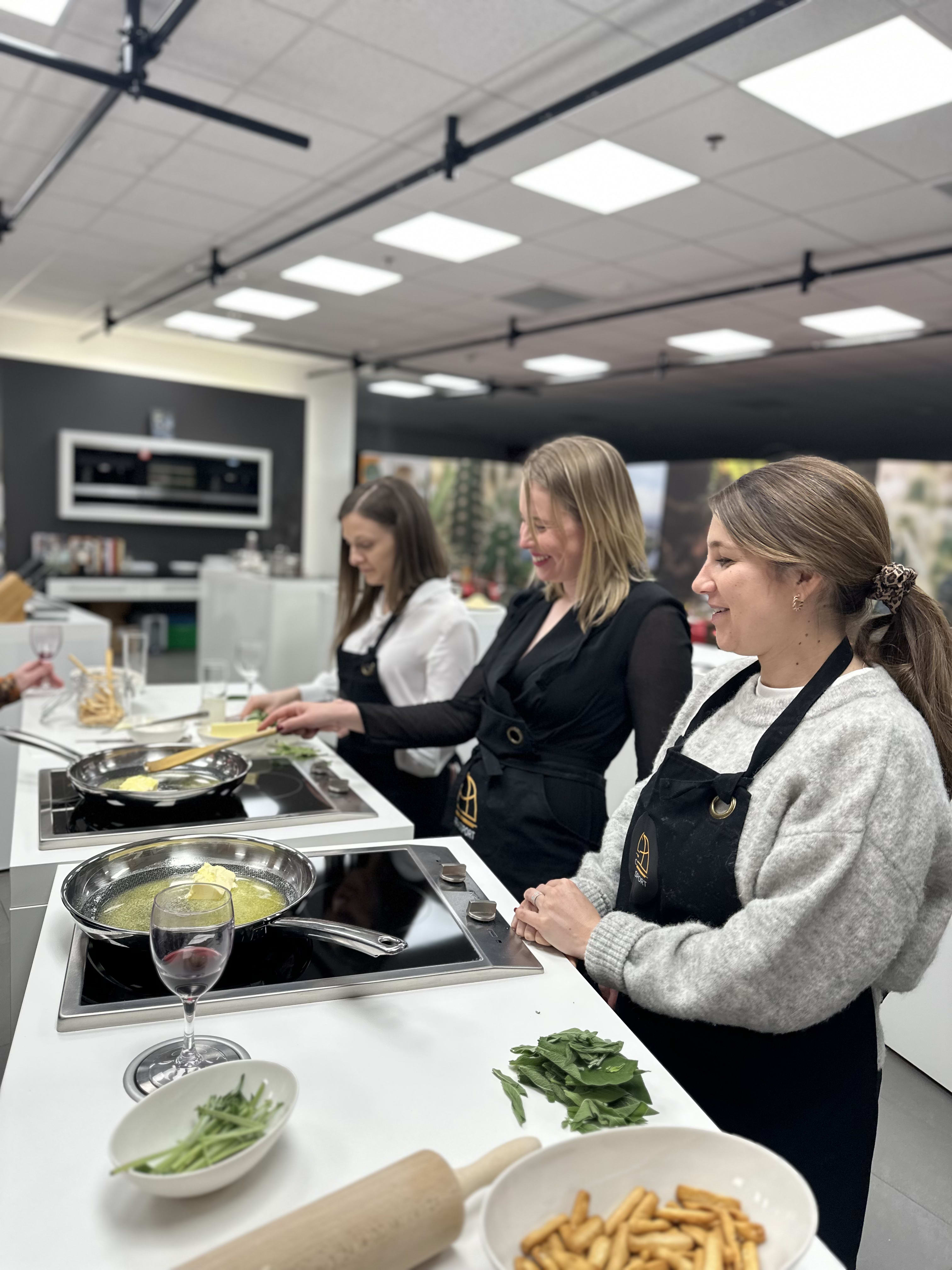 Three women in aprons cooking at stovetops during a culinary class in modern kitchen