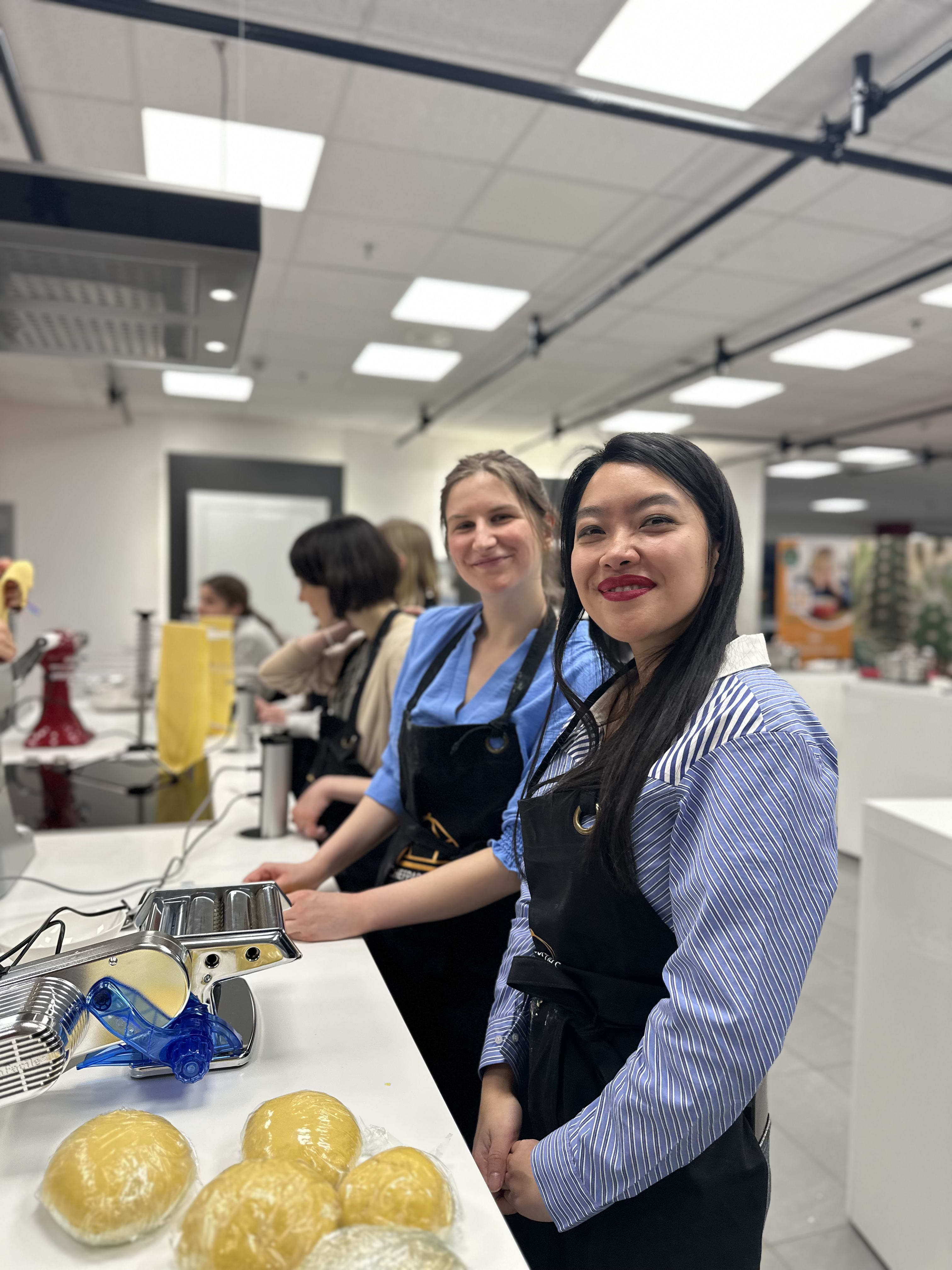 Two women in aprons smiling at camera during pasta-making class in commercial kitchen