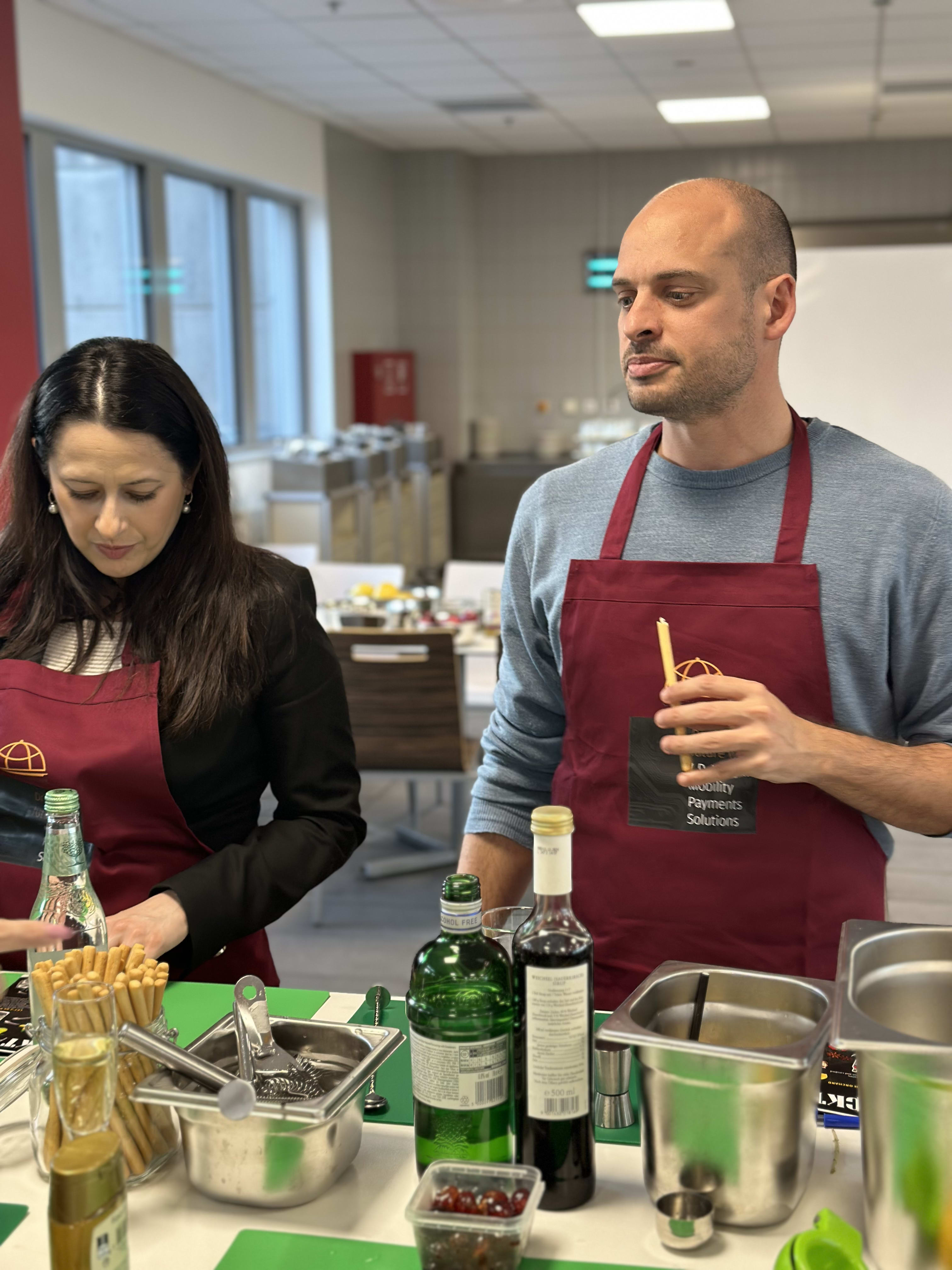 Two people wearing burgundy aprons preparing food and drinks at a cooking class station