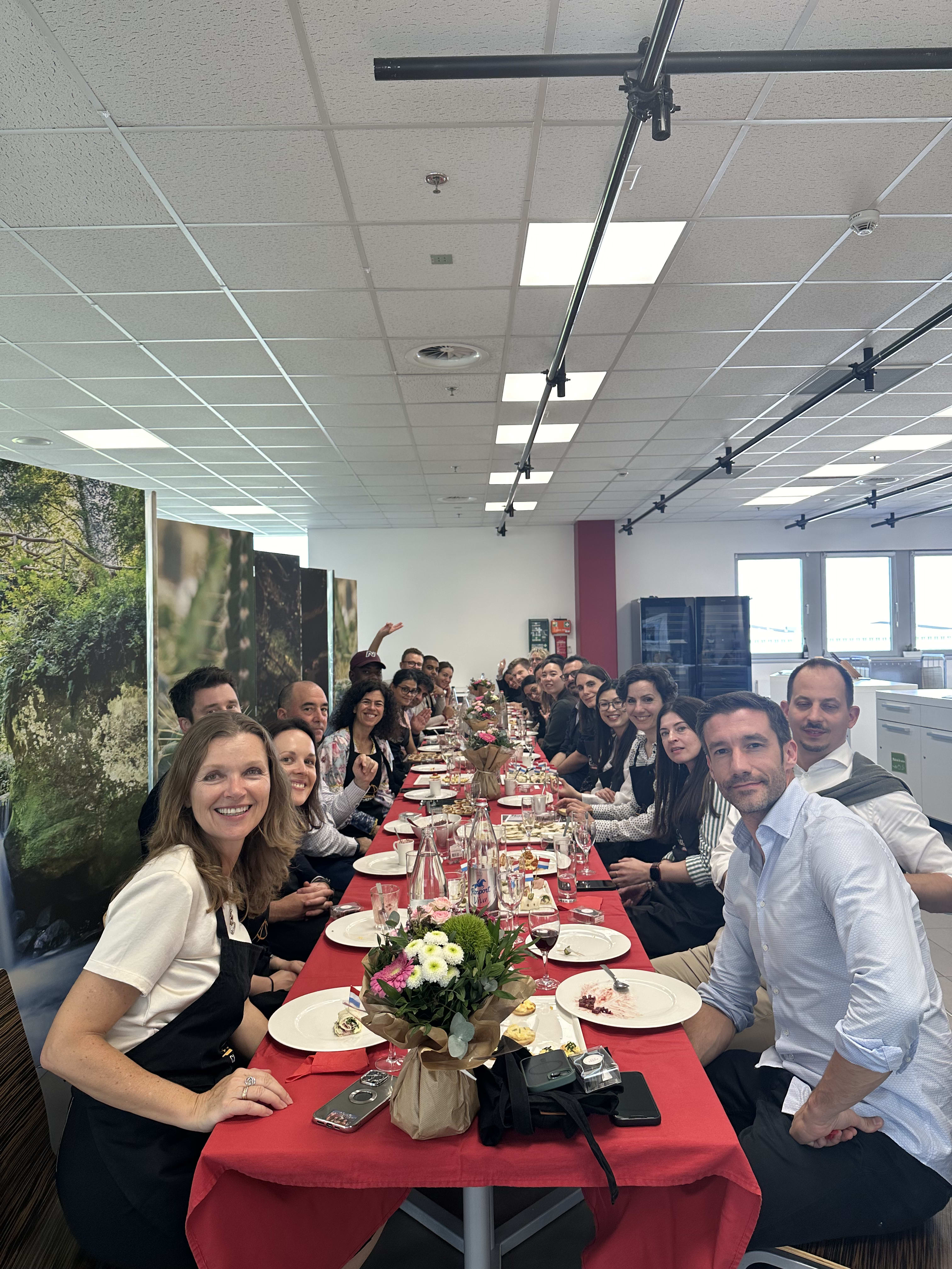 Large group of coworkers gathered around a long red table for an office celebration meal