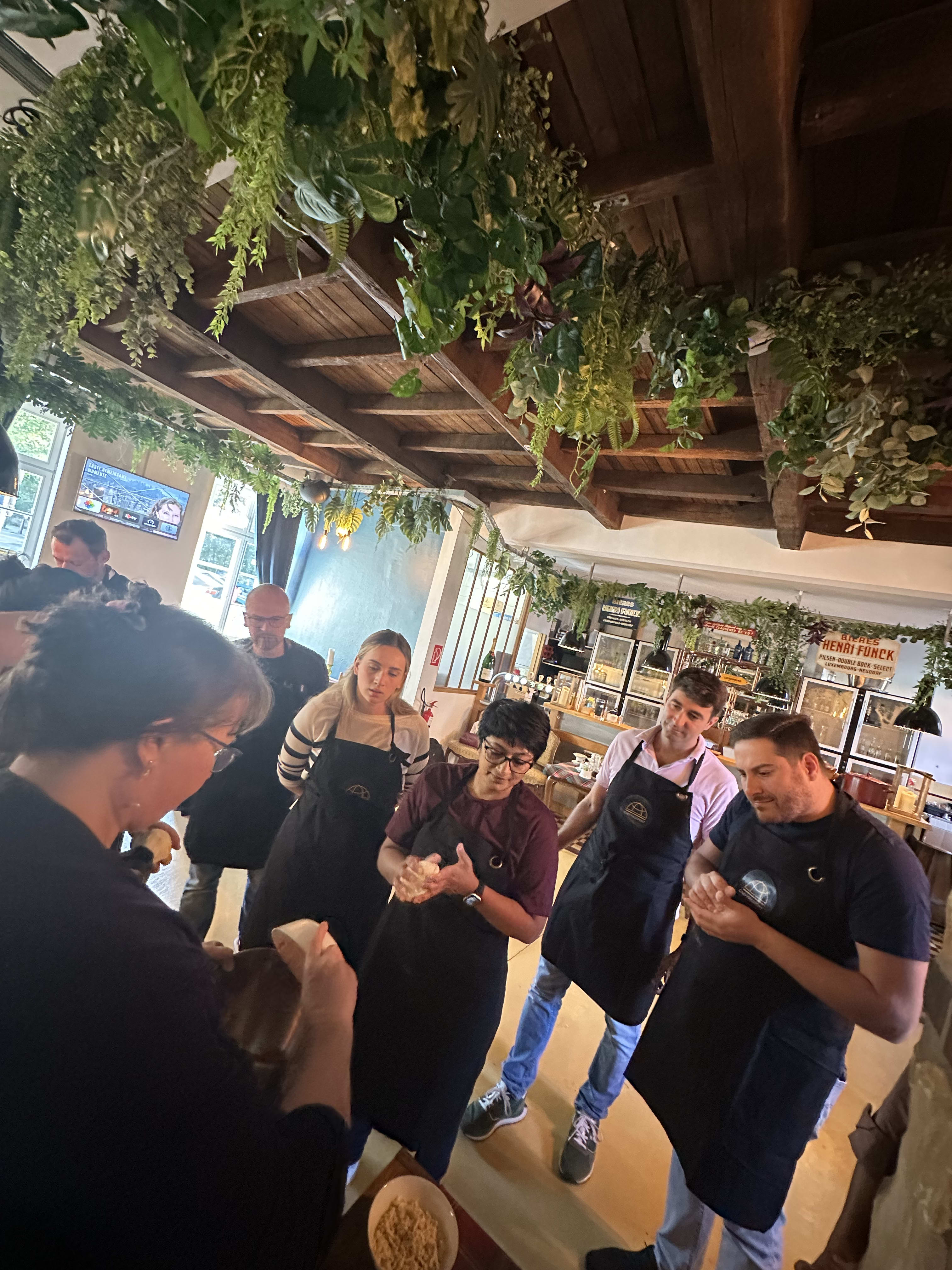 Group of people in black aprons attending a cooking class in restaurant with hanging plants