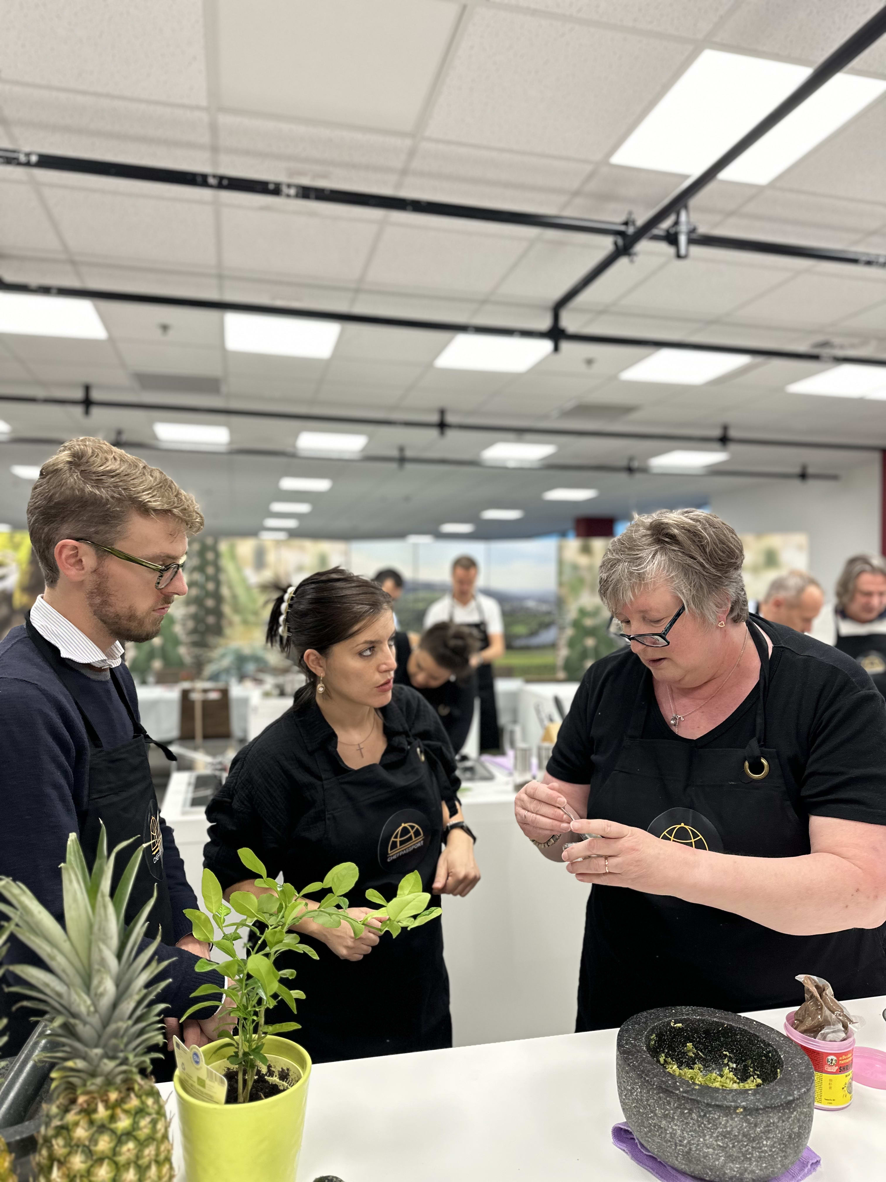 Three people in black aprons learning culinary techniques in a commercial kitchen classroom