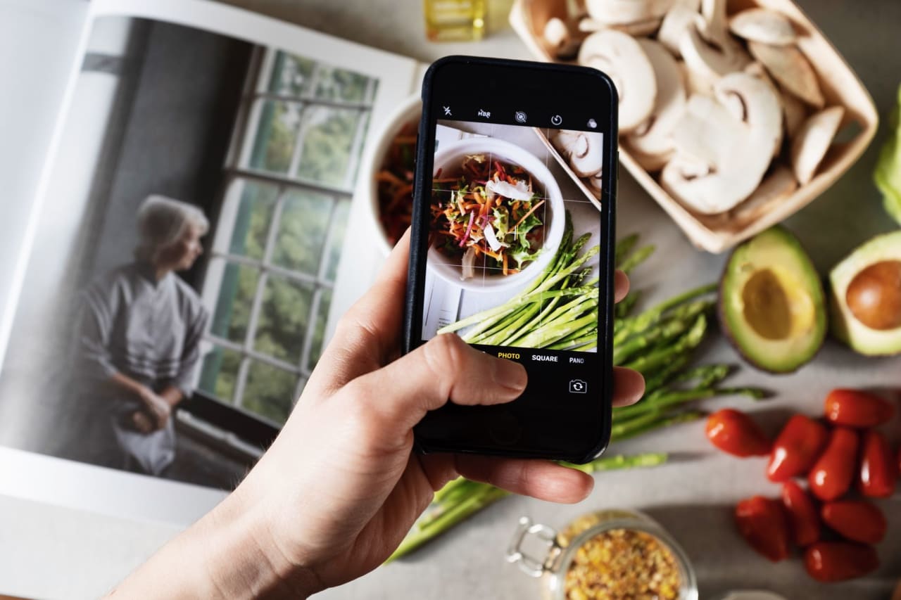 From above view of faceless blogger taking photo on smartphone of fresh tasty salad in white bowl and food placed around on table in modern kitchen