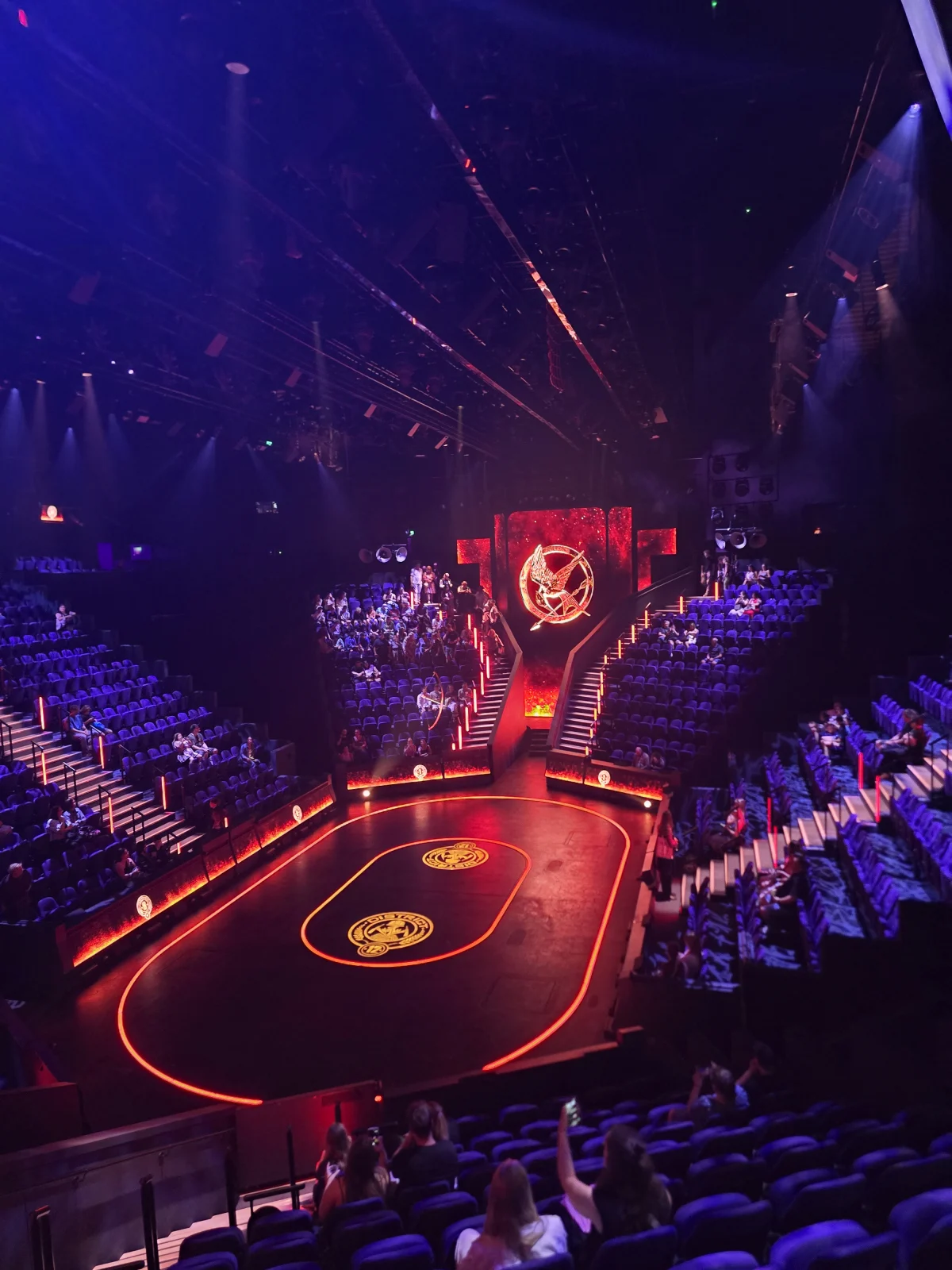 Wide view of The Hunger Games: On Stage arena in the round, with a glowing red Mockingjay symbol above the central staircase