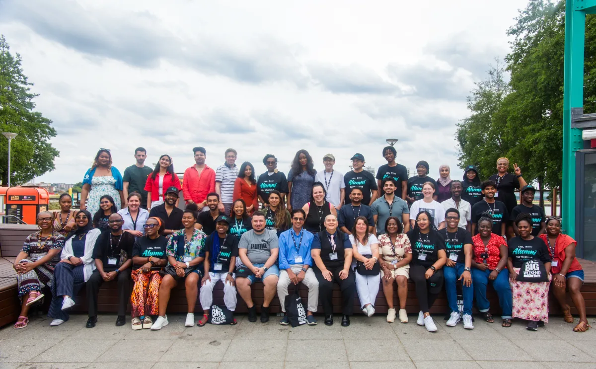 A large group photo of alumni standing and sitting in three rows outdoors, many wearing black 'UEL Alumni Big Weekend' t-shirts and lanyards.