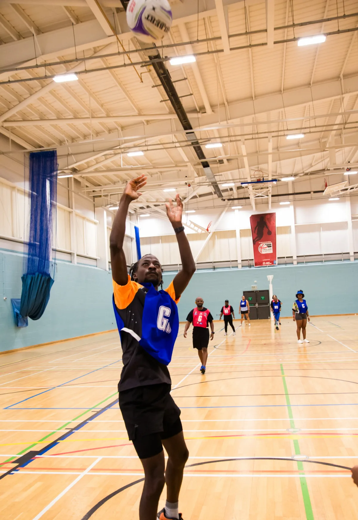 A man in a bib marked 'GS' shooting a netball mid-air on an indoor sports court.