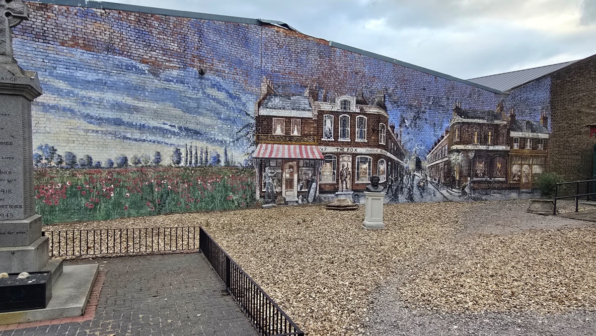 A large mural of a vintage London street scene with a poppy field, painted on a brick wall beside a war memorial.