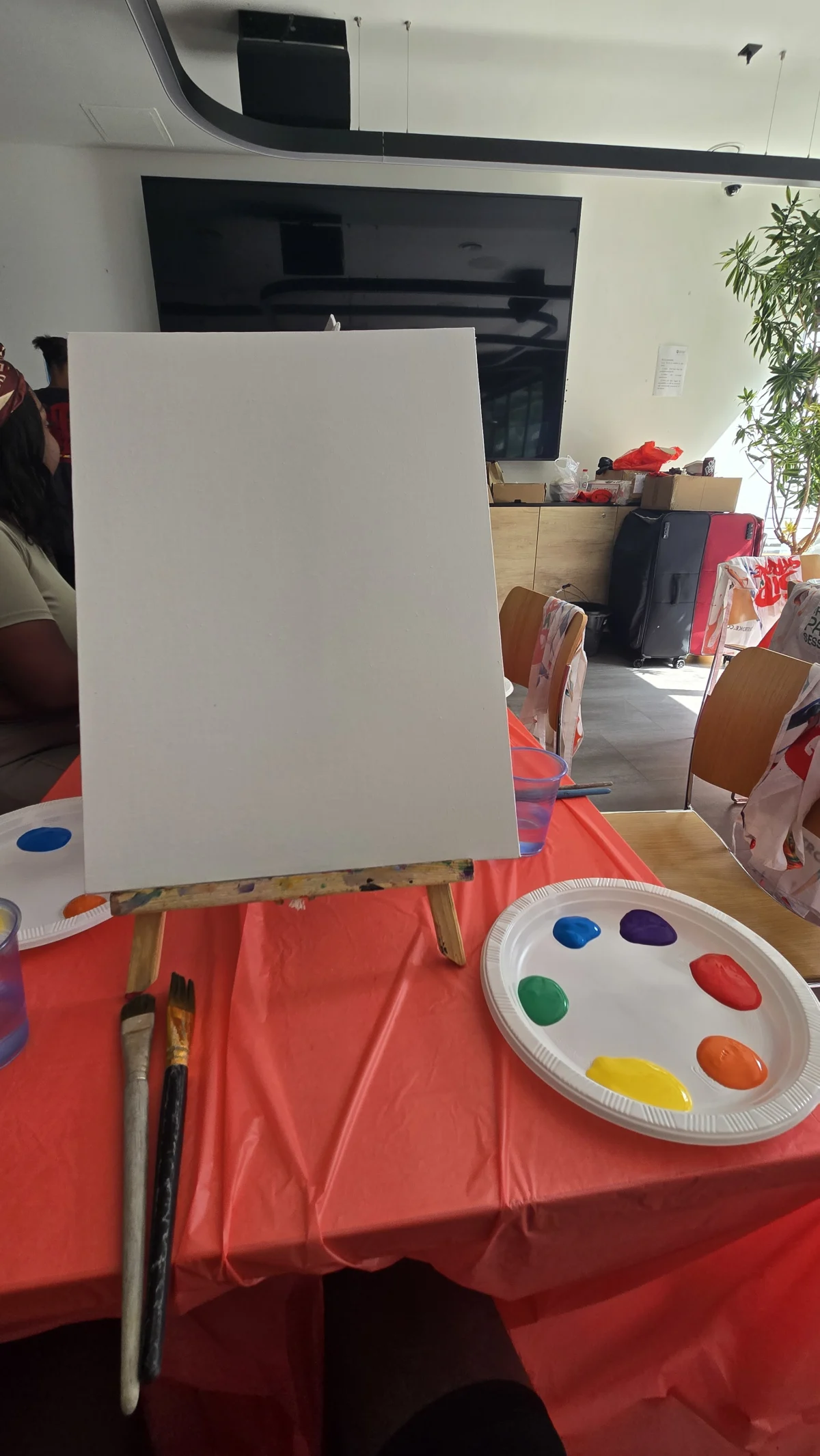 A blank canvas on an easel next to a paint-loaded palette and brushes, set up for the Sip and Stroke painting session.