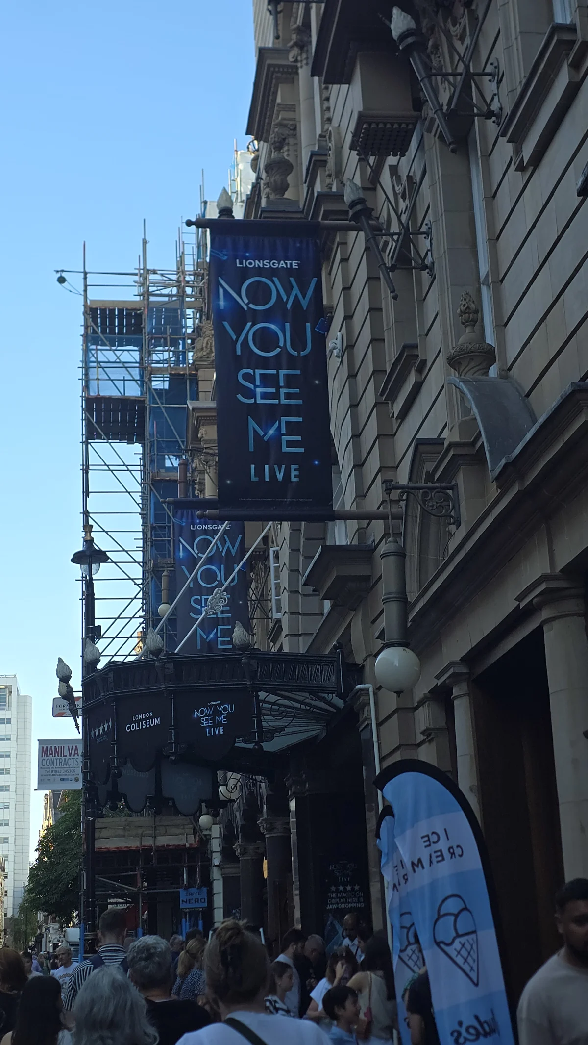 The London Coliseum's exterior with two 'Lionsgate: Now You See Me Live' banners hanging above the entrance, a crowd gathered below.