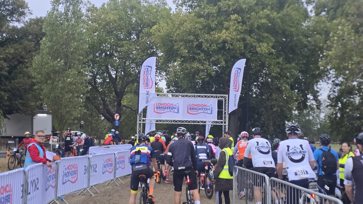 Cyclists gathered at the start line beneath a London to Brighton Cycle Ride banner arch, early morning with marshals in hi-vis directing the queue.