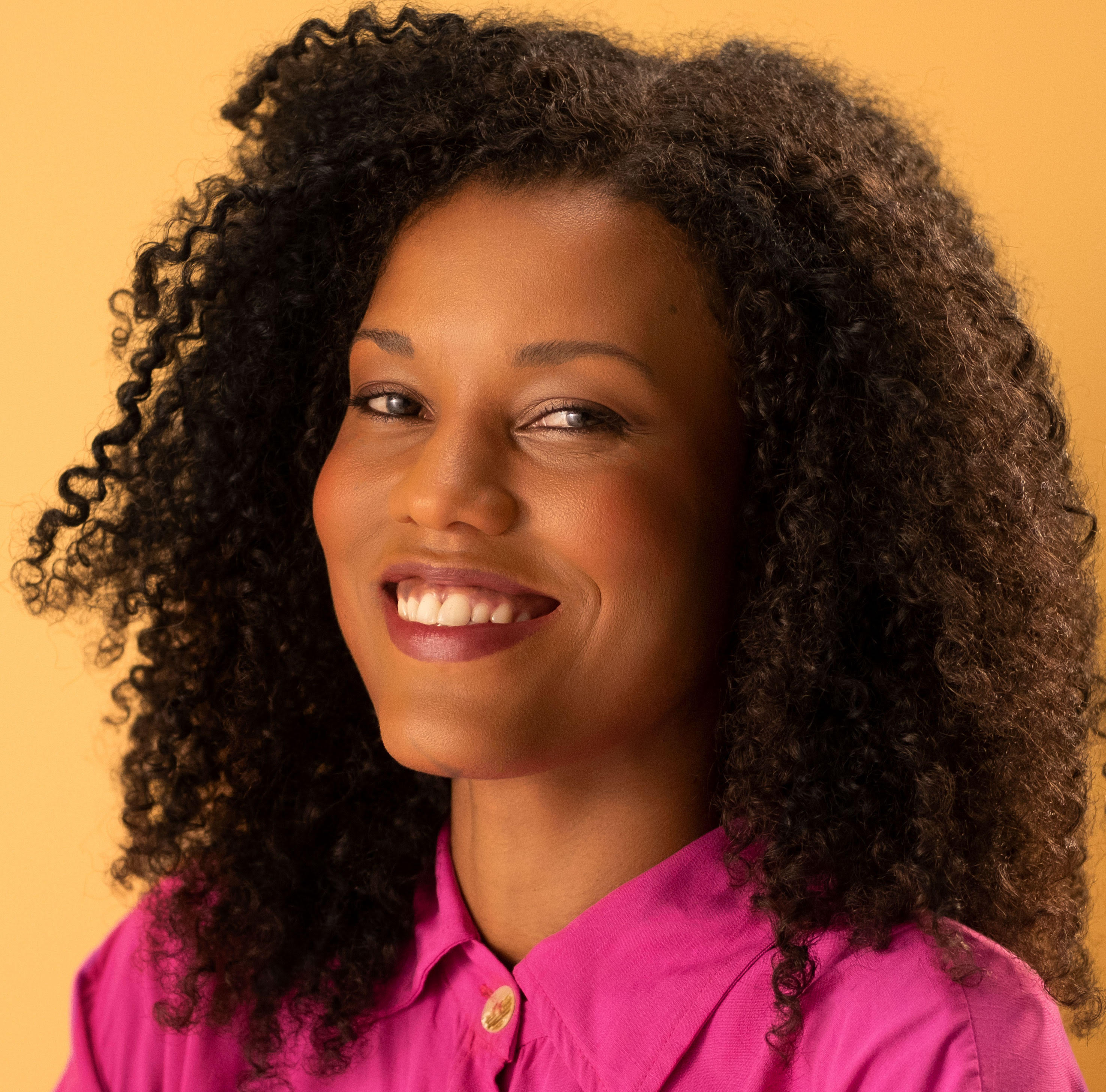 foto de rosto de Wanuzia Braga, mulher negra sorrindo, com cabelos cacheados soltos na altura do ombro, veste blusa sem manga laranja.