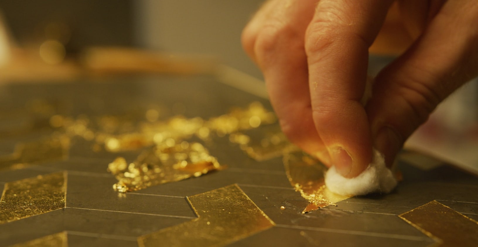 A hand burnishing squares of 24k gold leaf onto the reverse of a dark glass panel with a cotton pad, the leaf laid edge to edge in a grid