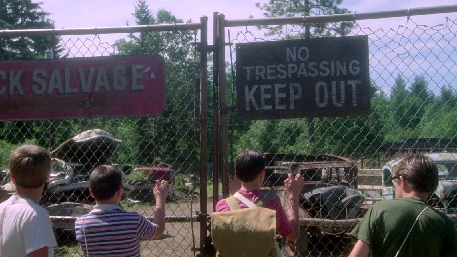 four boys standing at a fence looking into a Junkyard