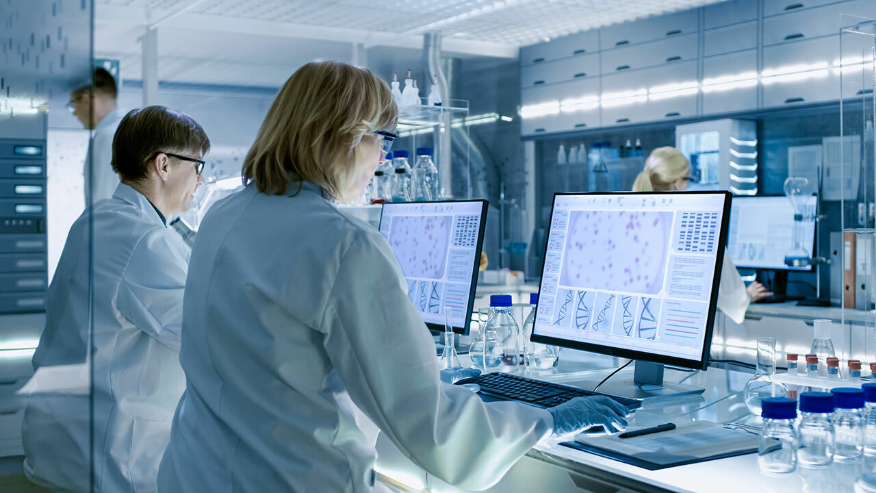 Two medical researchers in white lab coats sit at a lab bench examining microscopic images on two large computer monitors