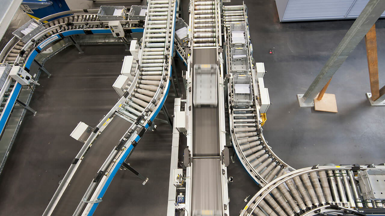 A conveyor belt system in a warehouse or manufacturing facility, featuring multiple roller belts with blue side rails curving around corners. The belts are supported by metal frames, with various mechanical components visible along the sides