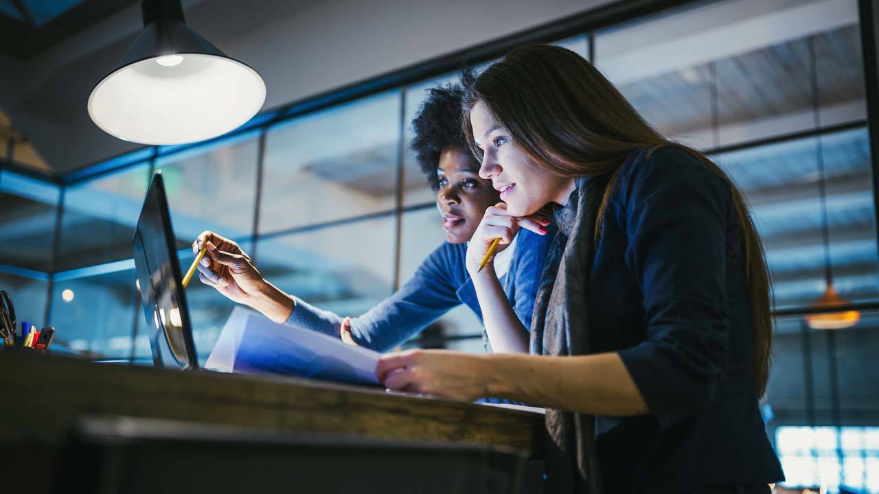 Two individuals at desk, one pointing at computer screen while the other holds documents