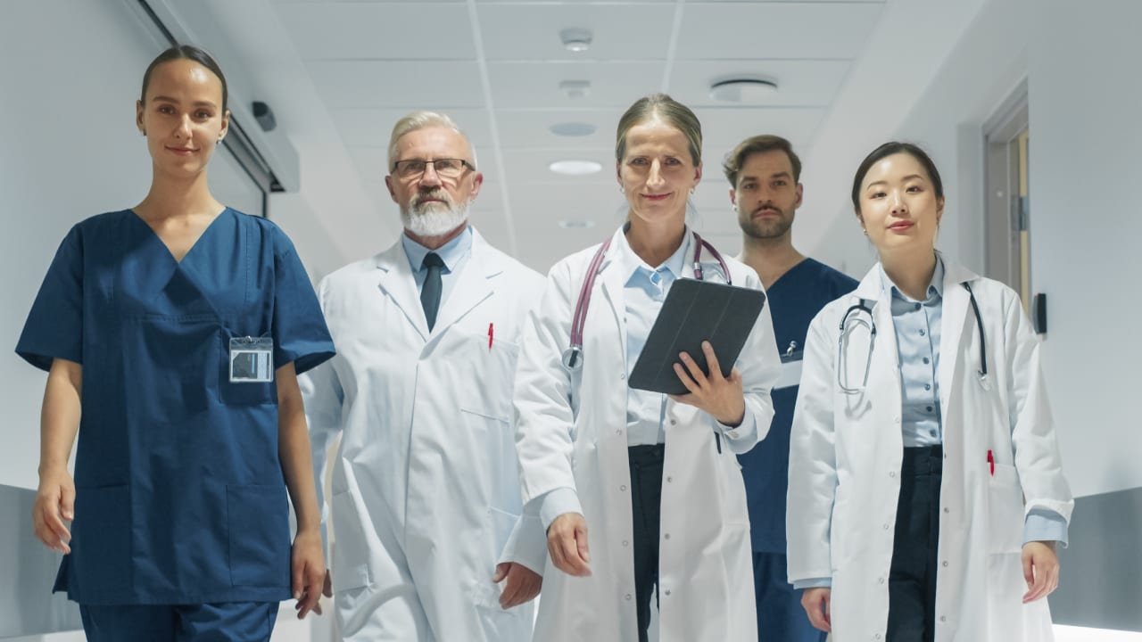 A group of five medical professionals walking down a hospital corridor. The group includes three in white lab coats, one in blue scrubs, and one in blue and white attire. One person is holding a clipboard, and two have stethoscopes around their necks. The corridor is well-lit with ceiling lights and doors on either side