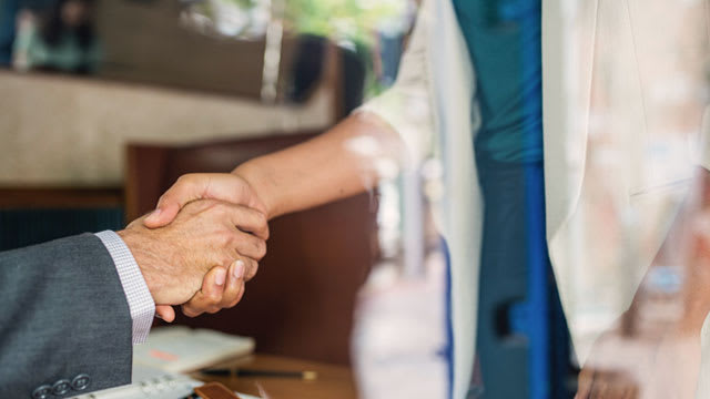 Two people shaking hands, one wearing a suit and the other in casual attire, with a blurred background of an office setting