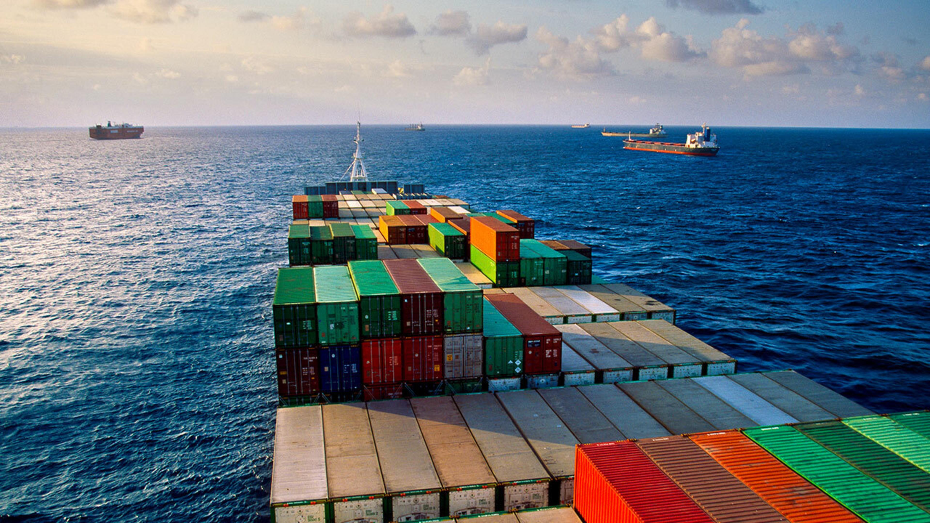 A large cargo ship sailing on the ocean, carrying numerous colorful shipping containers stacked in rows. The containers are green, red, blue, yellow, and orange. The ship's deck is visible in the foreground, with additional ships on the horizon. The sky is clear and the sea is calm