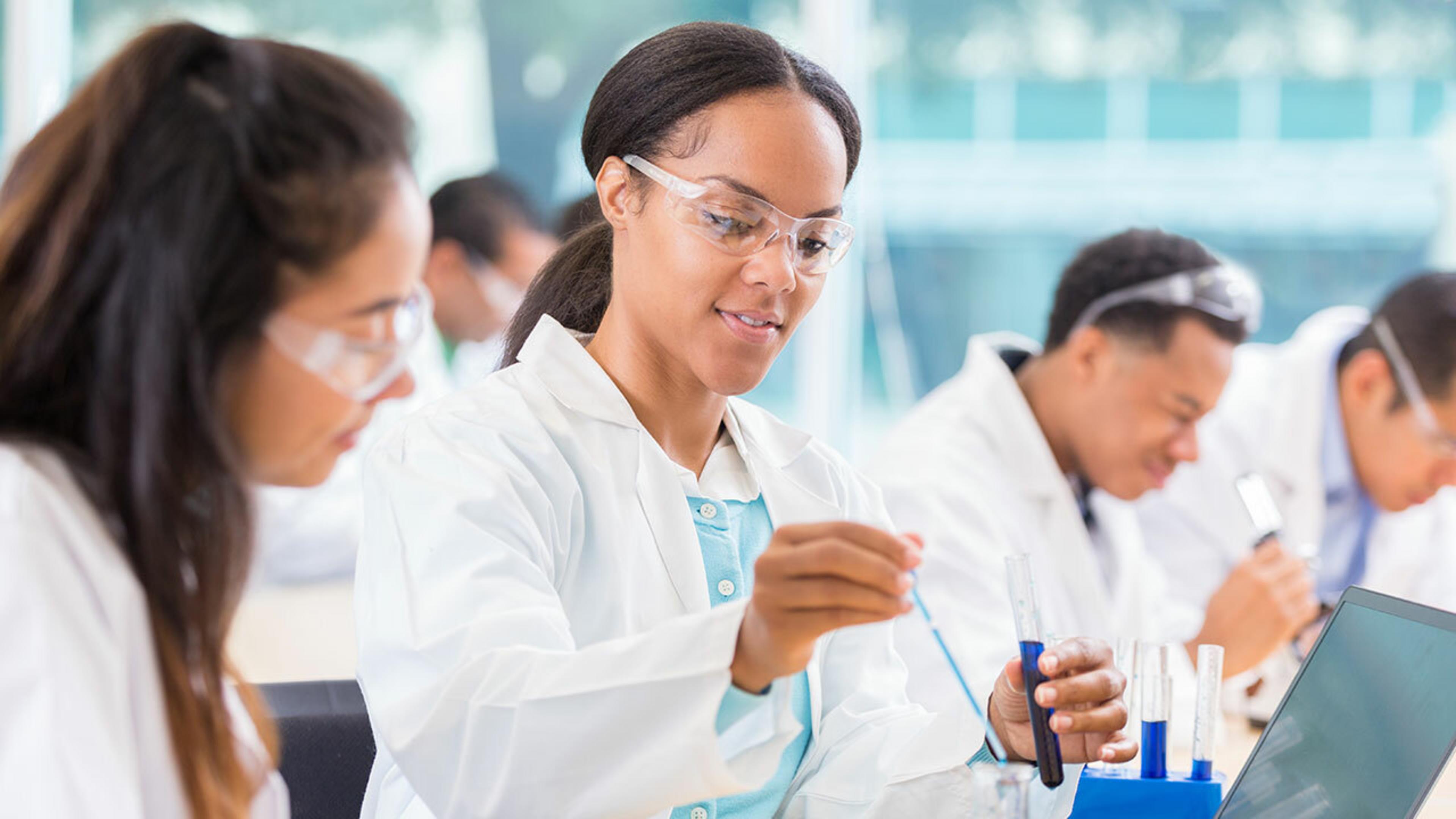 A group of medical professionals working in a laboratory, wearing lab coats and safety glasses, using a pipette to transfer liquids between test tubes