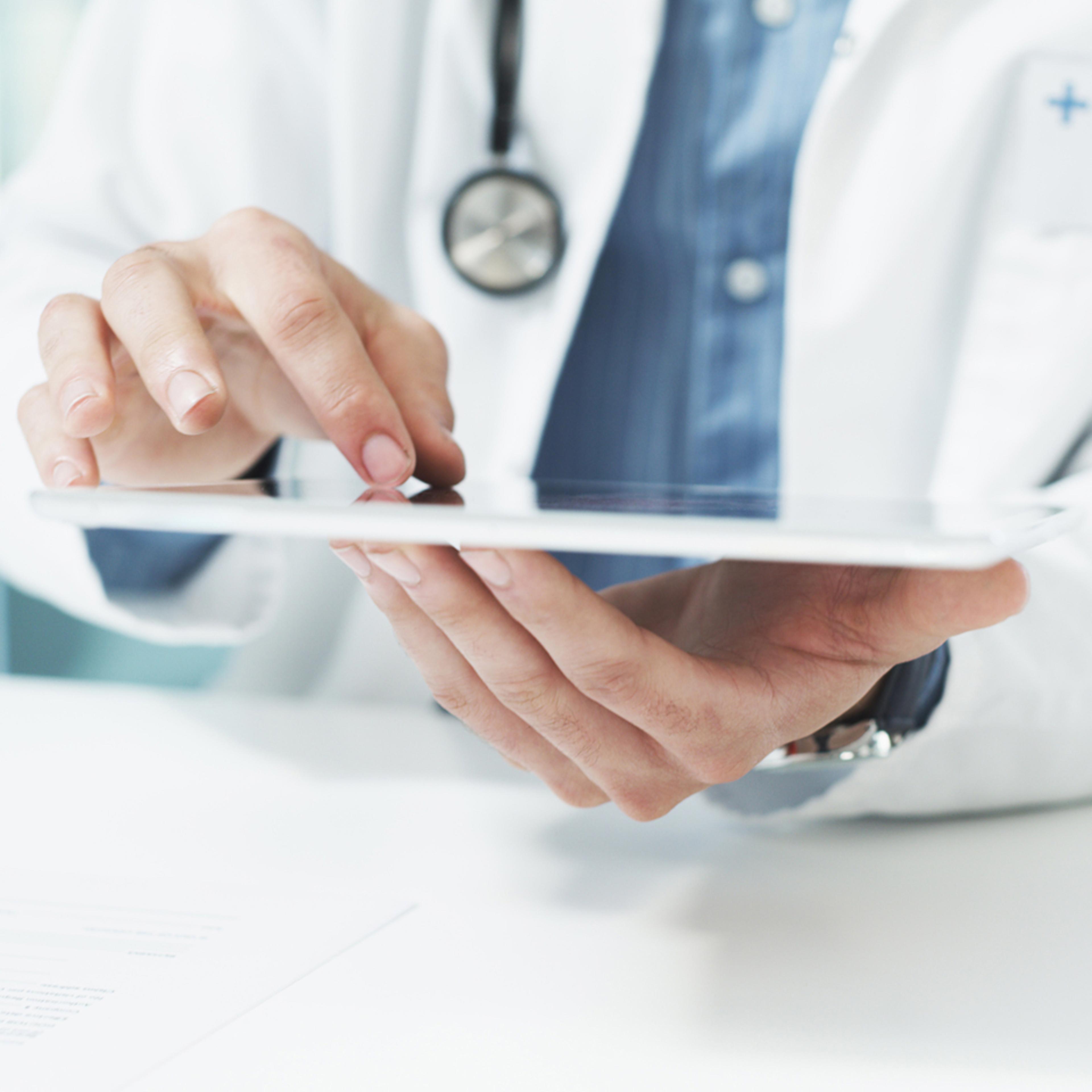 A medical professional wearing a physician's coat and stethoscope interacts with an app on a tablet device