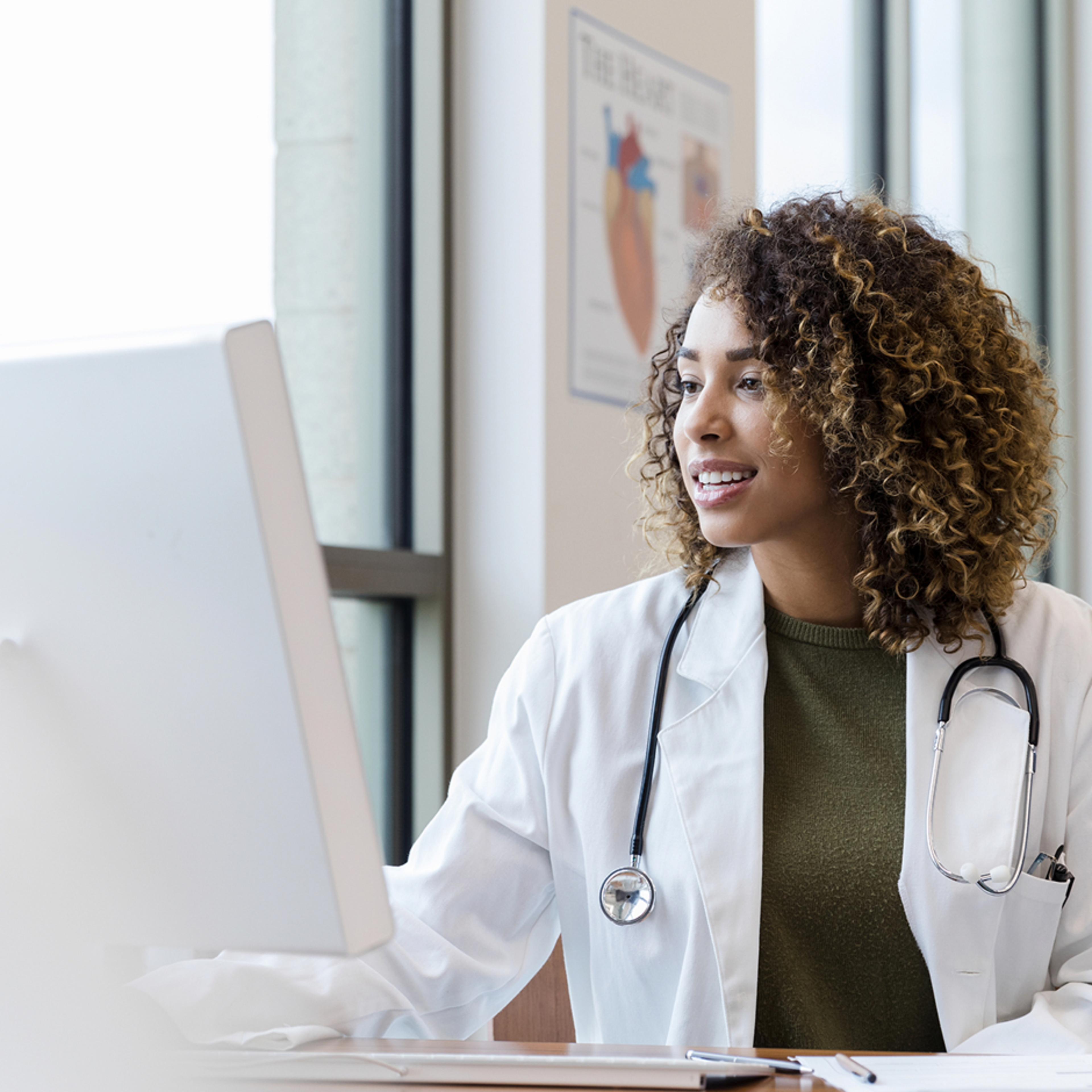 A medical professional with curly hair, wearing a white lab coat and stethoscope, is seated at a desk in front of a computer. A poster of the human heart is visible on the wall in the background
