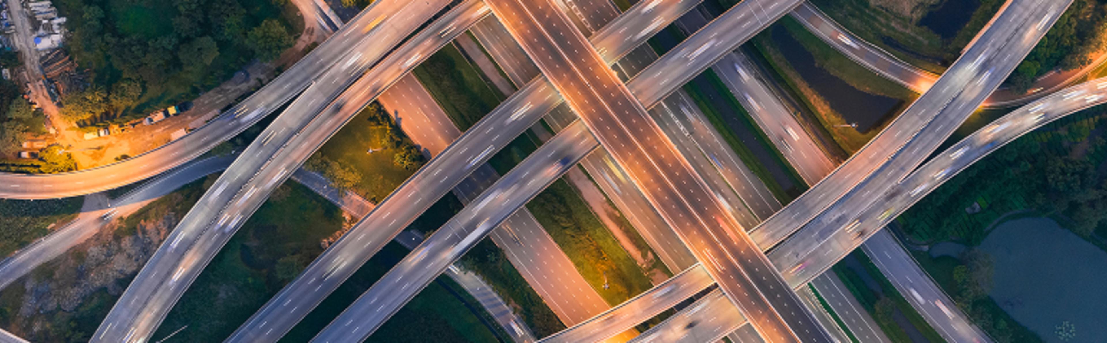An aerial view of a complex highway interchange with multiple layers of intersecting roads, illuminated by streetlights and light trails from moving vehicles. The surrounding area features patches of greenery and water bodies