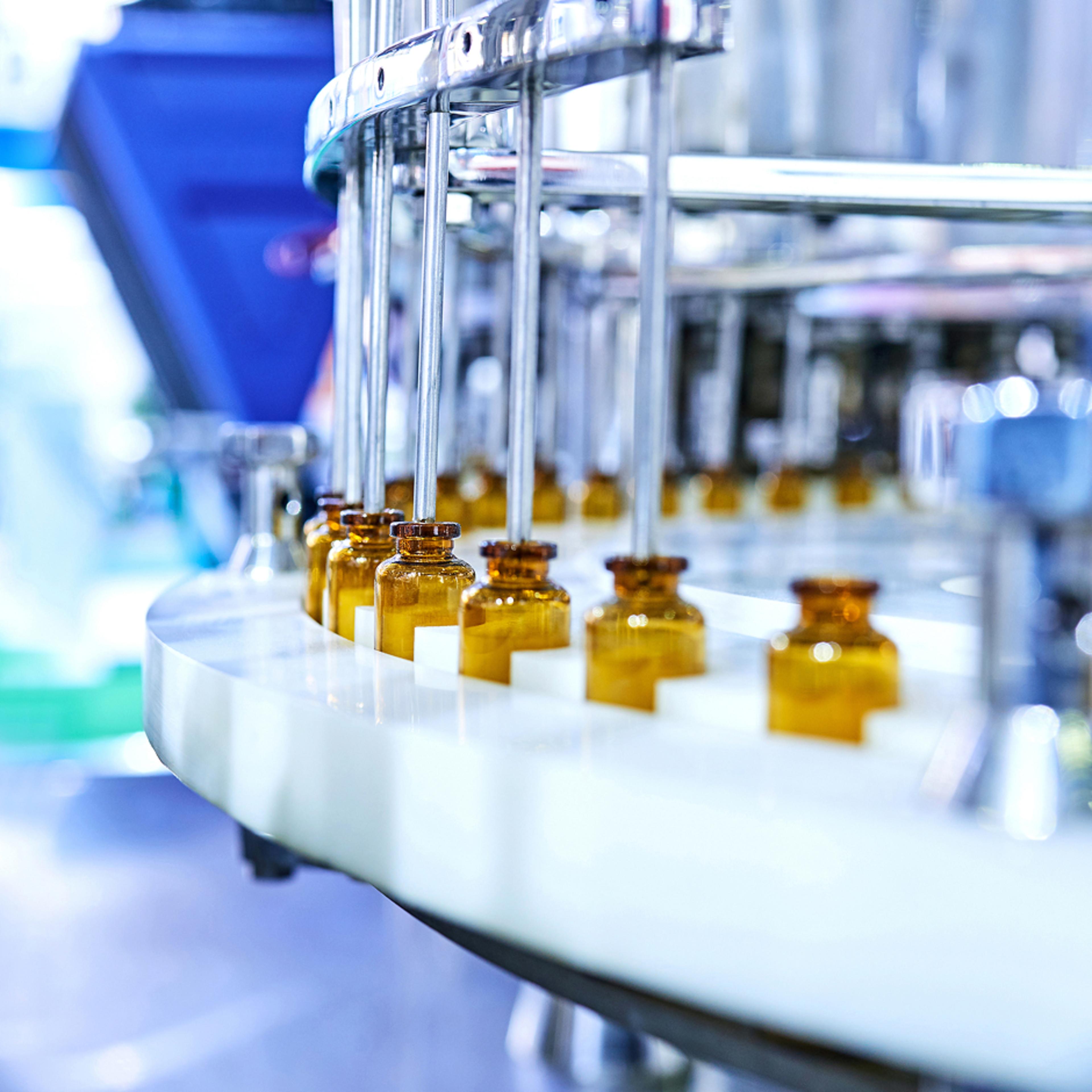 A close-up view of a pharmaceutical production line with multiple brown glass vials being filled by automated machinery. The vials are arranged in a row on a conveyor belt, with metal rods dispensing liquid into each vial. Industrial equipment is visible in the background