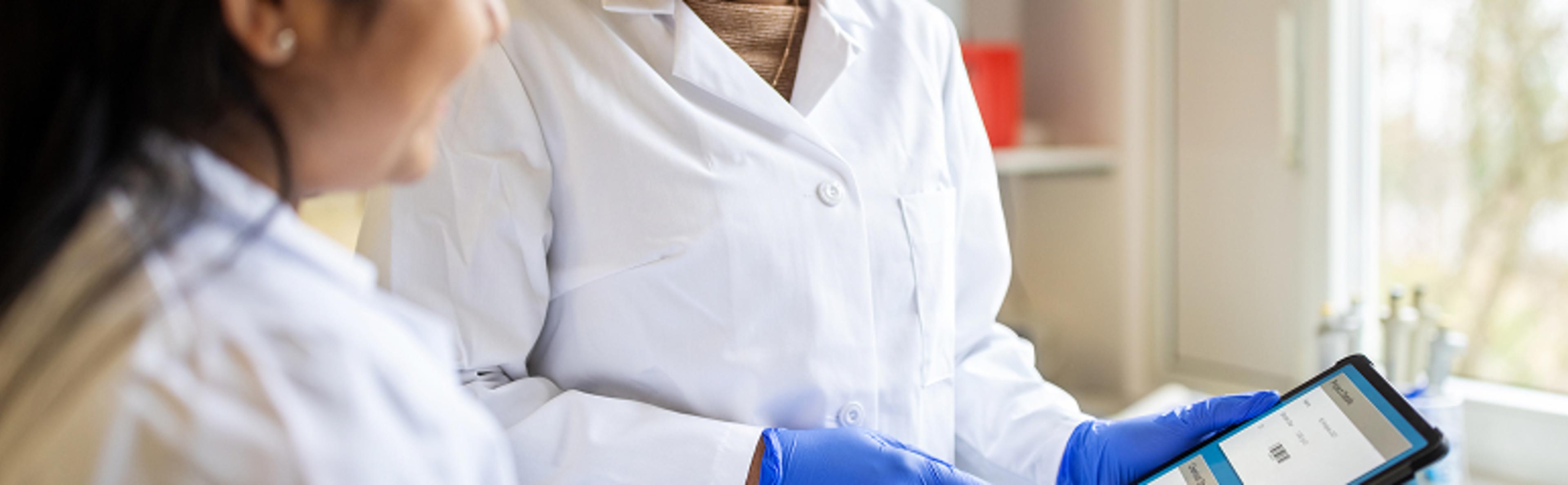 Two healthcare professionals in a lab. One is smiling while the other, wearing blue gloves, is holding a tablet with some form of data displayed