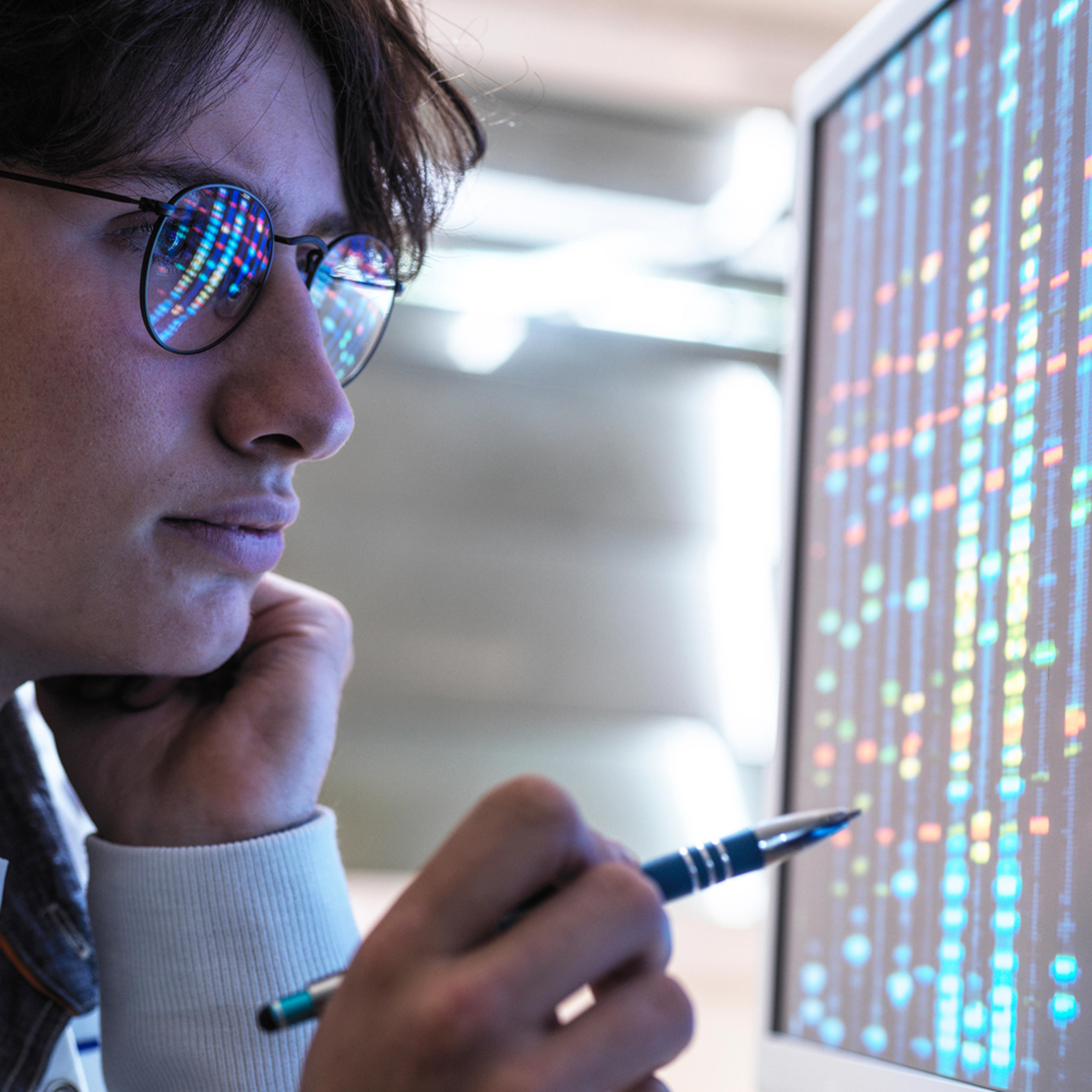 A close-up of a medical professional wearing eyeglasses and gazing thoughtfully at a computer screen, holding a pen