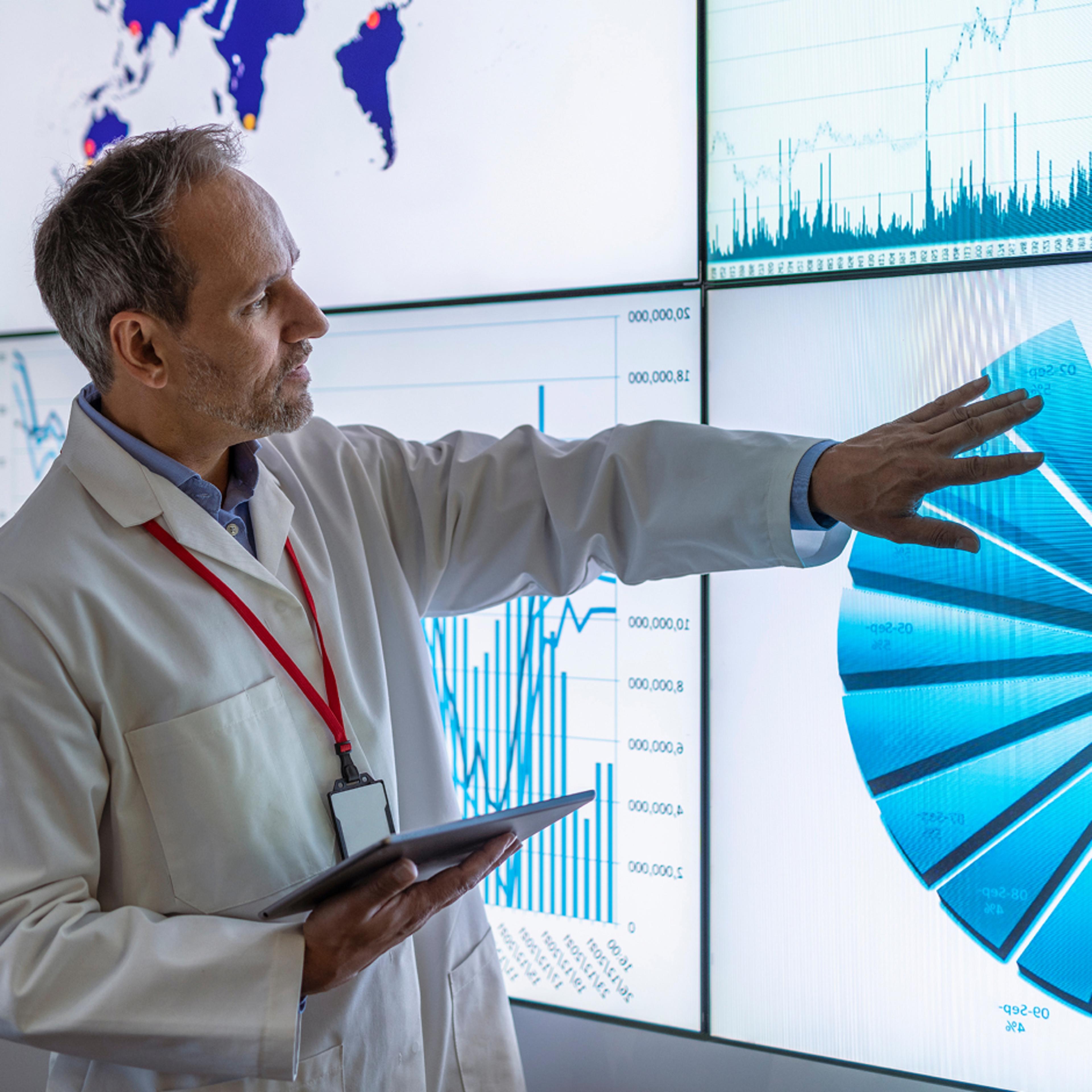 Lab coat-wearing figure points at charts on screens while holding a tablet