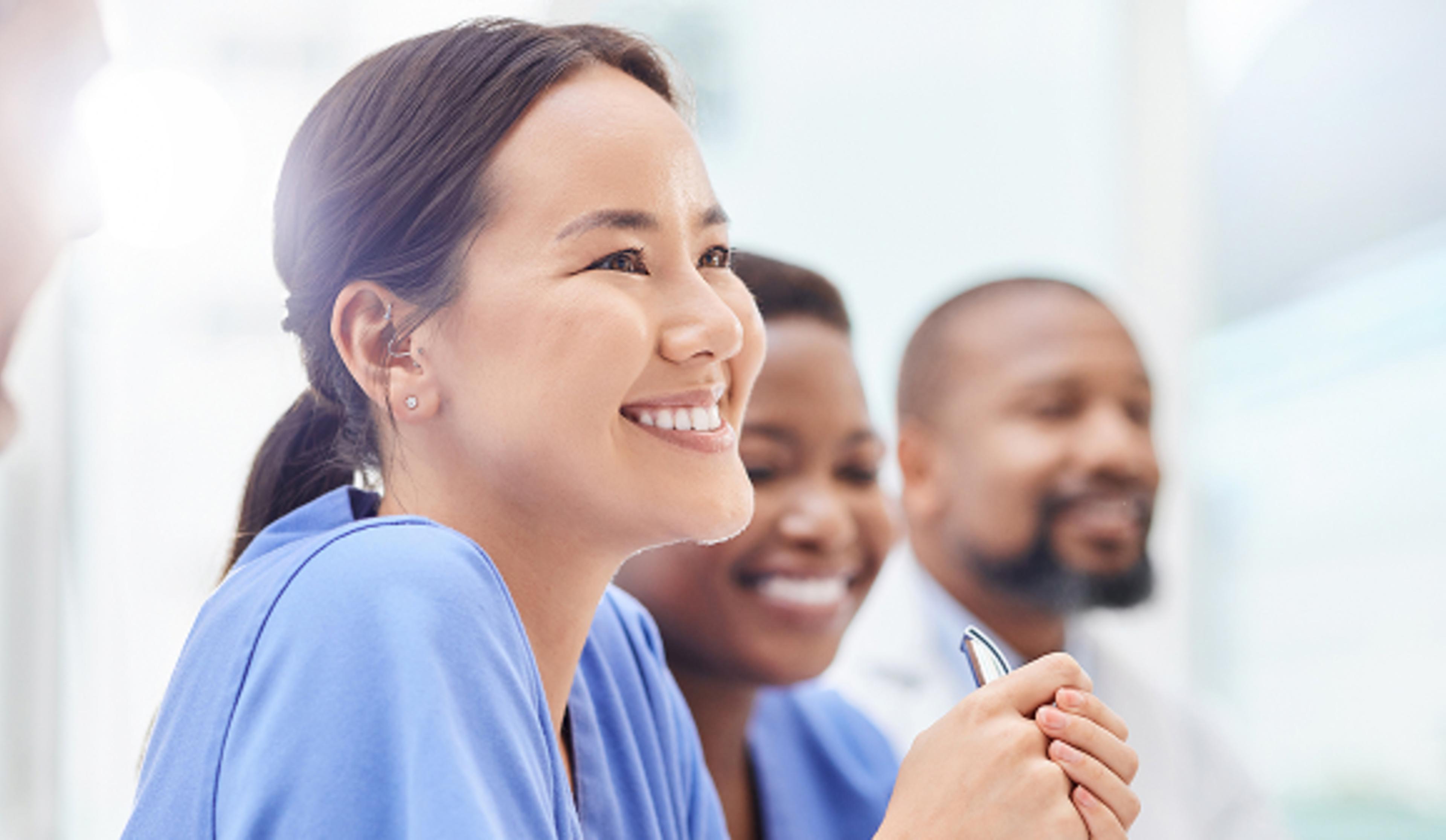 A group of medical professionals in blue scrubs are seated and engaged in a discussion. One person is holding a pen, indicating active participation. The bright, out-of-focus background suggests a well-lit room or hospital setting