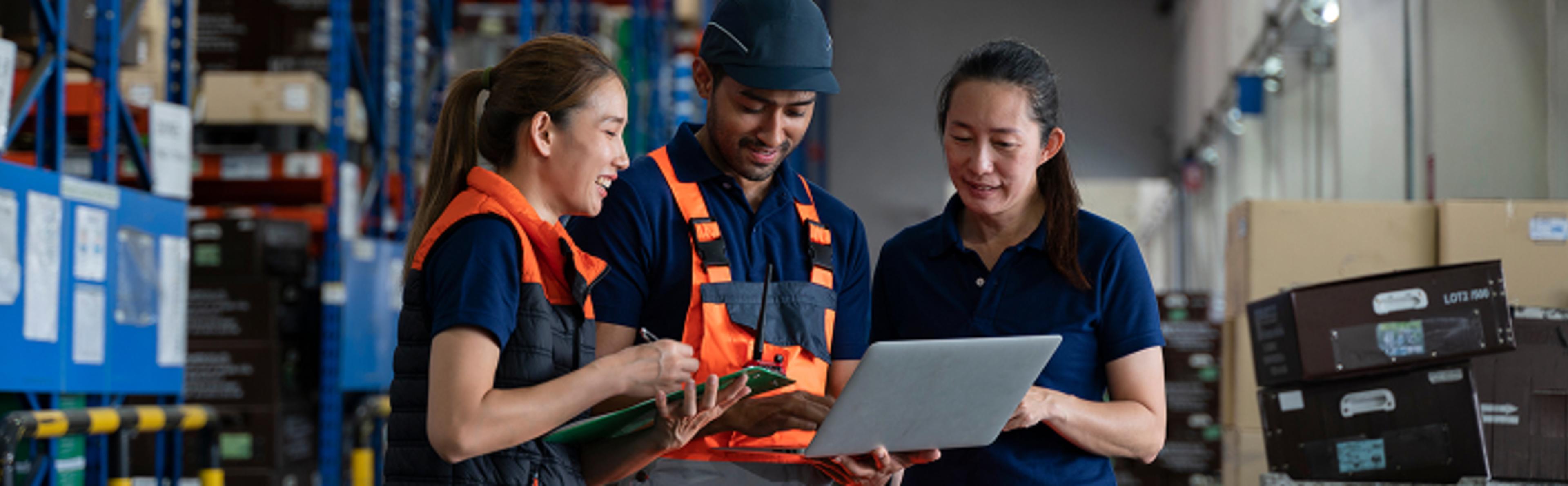 Three warehouse workers are standing together in a storage area. Two are wearing orange safety vests and holding a clipboard, while the third is holding a laptop. Shelves with various items and boxes are visible in the background