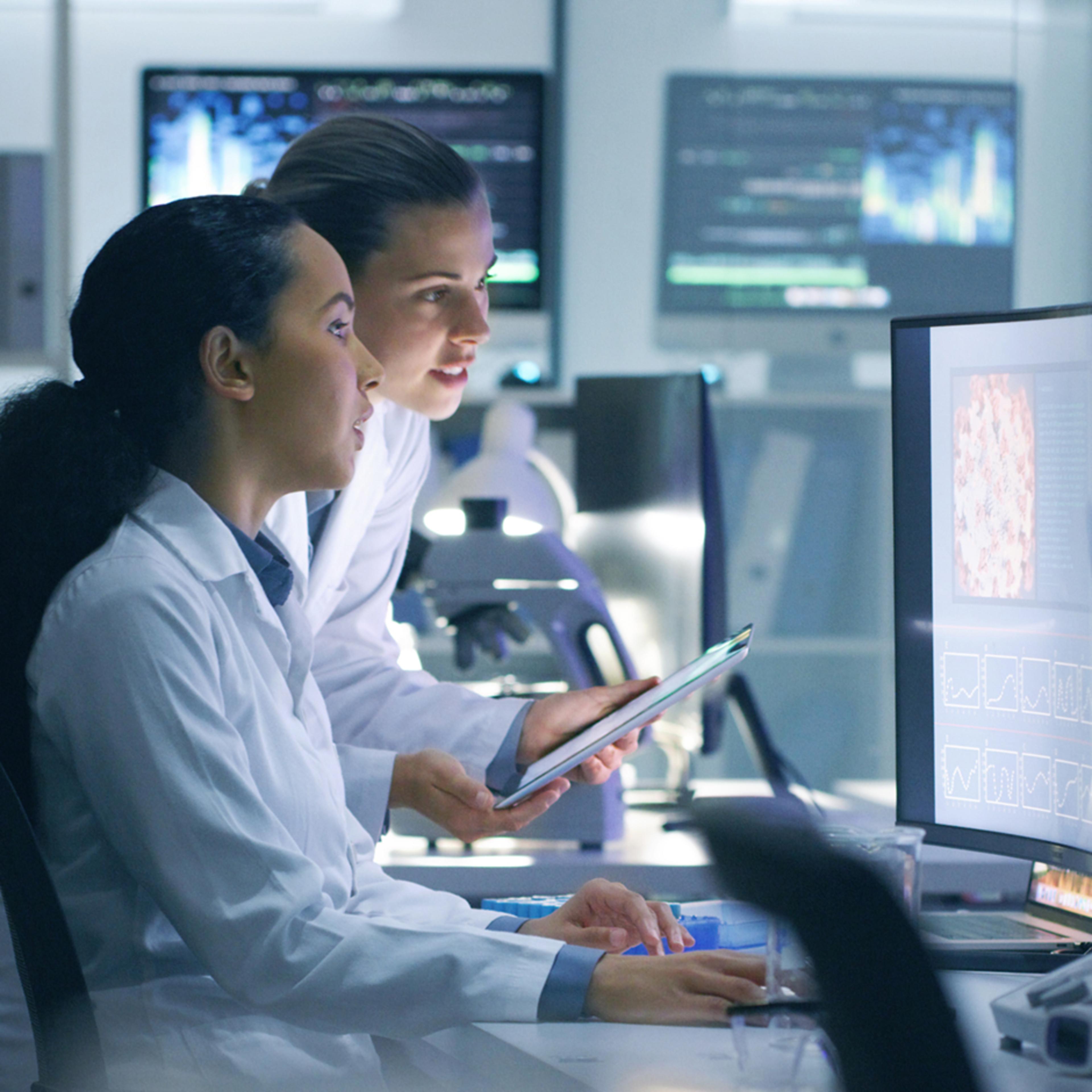 Two scientists collaborate on a project, inspecting a tablet device and a computer monitor in a hi-tech lab