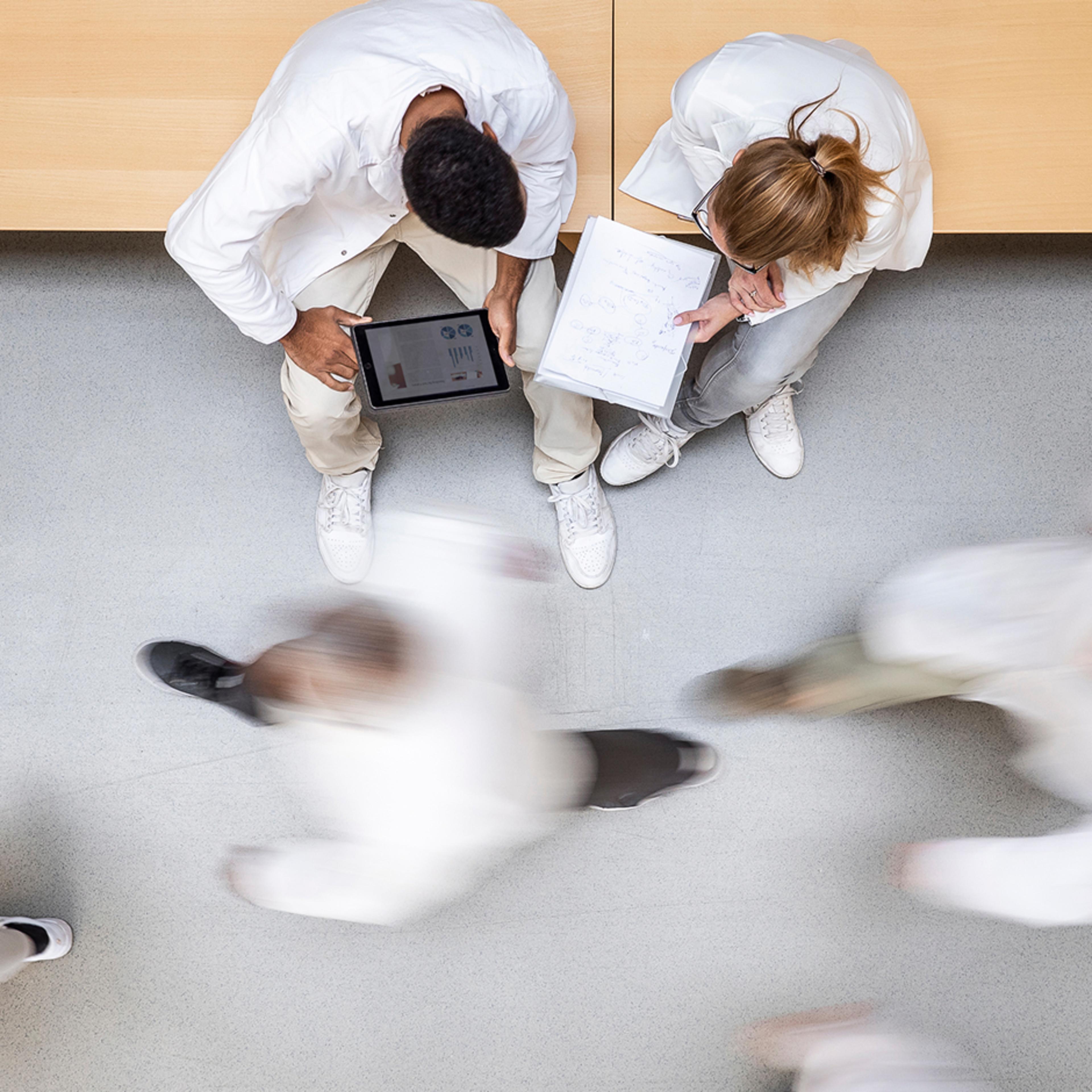 Two people in white coats on bench, one with tablet, one writing, with blurred figures passing by